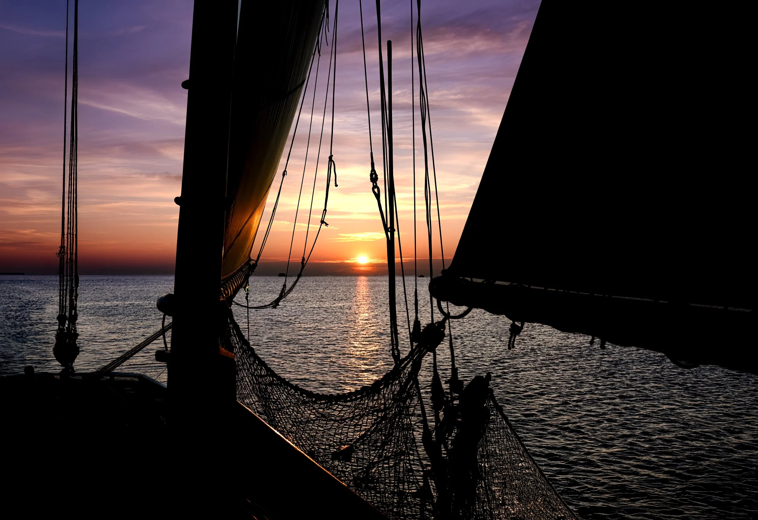 View of a sailboat's mast and rigging silhouetted against a colorful sunset over the ocean, with soft pink, purple, and orange clouds in the sky.