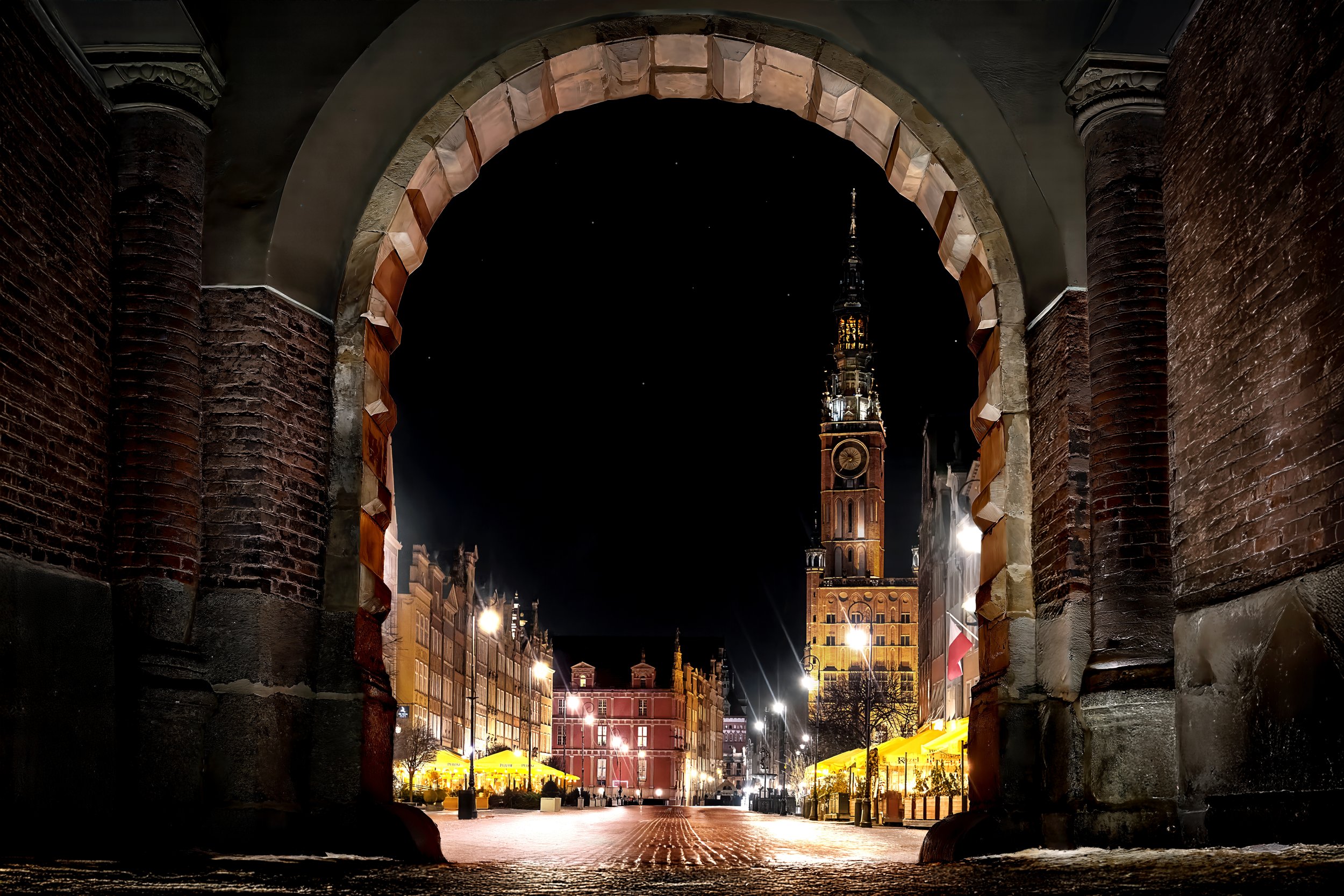 Night scene of a city street viewed through an ancient brick archway, with a tall illuminated tower or church spire in the background and yellow street market umbrellas in the foreground.