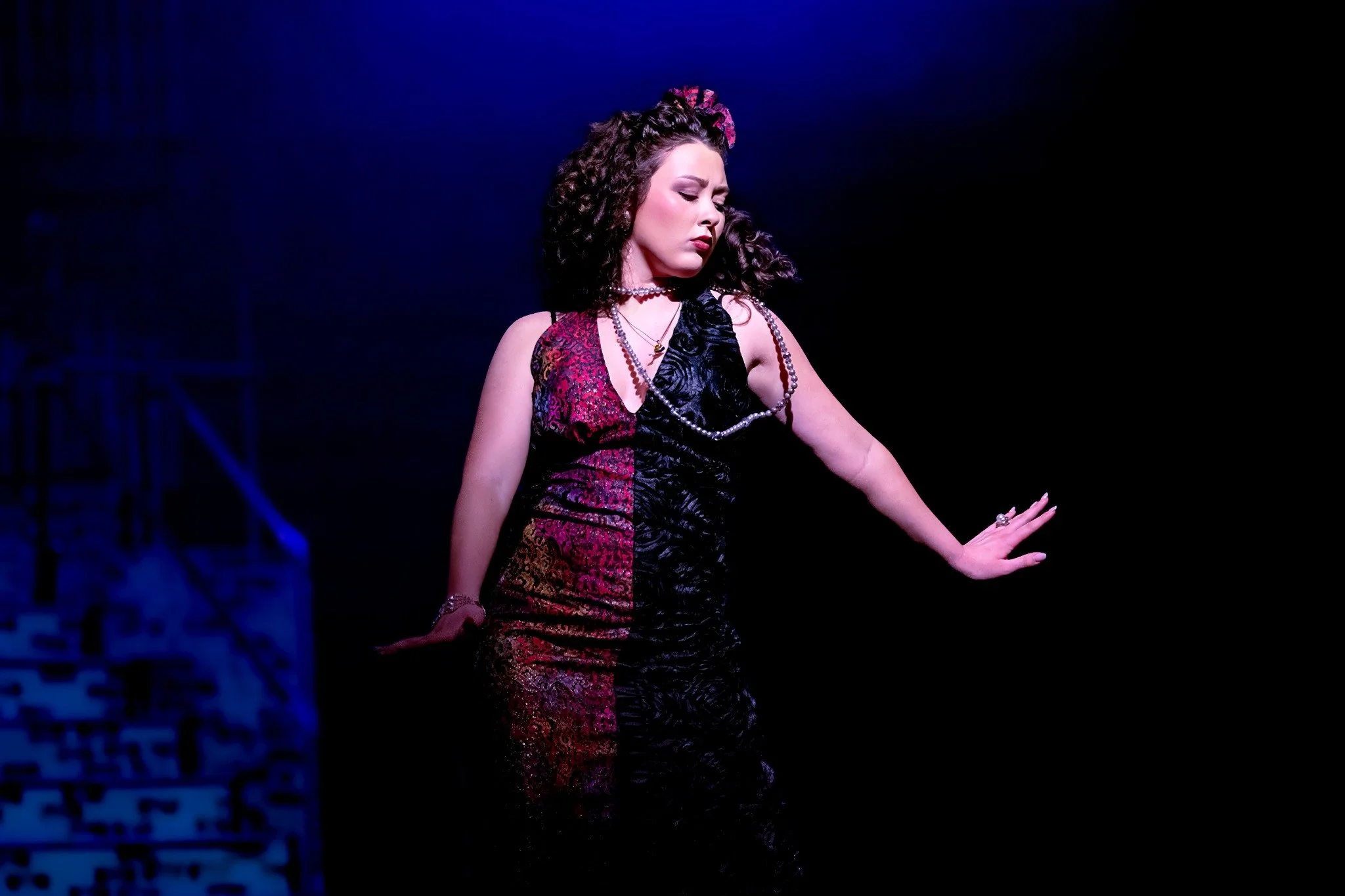 A woman with styled curly hair, dressed in a patterned dress, wearing jewelry, appears to be dancing or performing on stage with a dark background and subtle lighting.