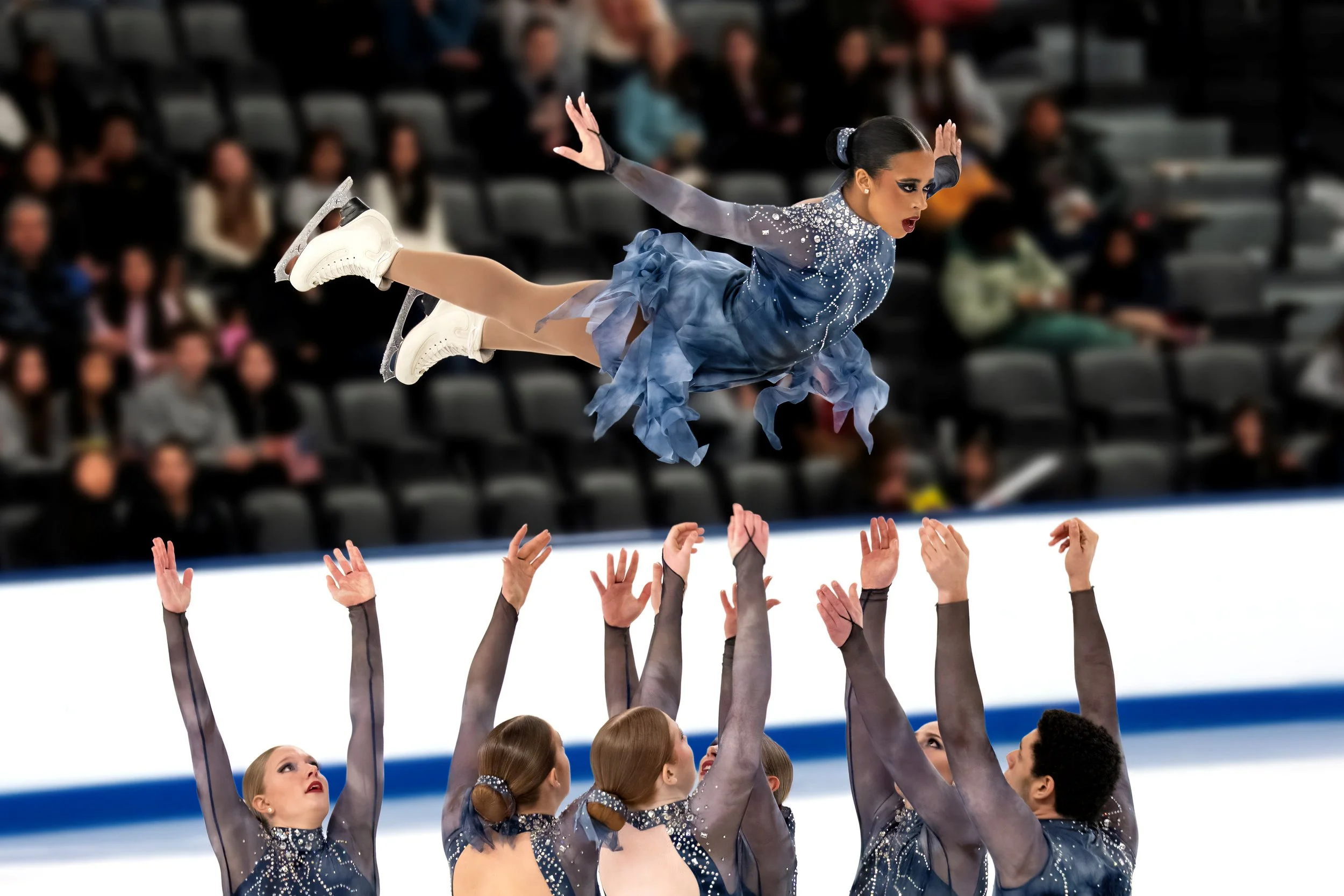 An ice skating team performing a lift in the air with a young female skater in a shimmering blue dress, with an audience watching in the background.