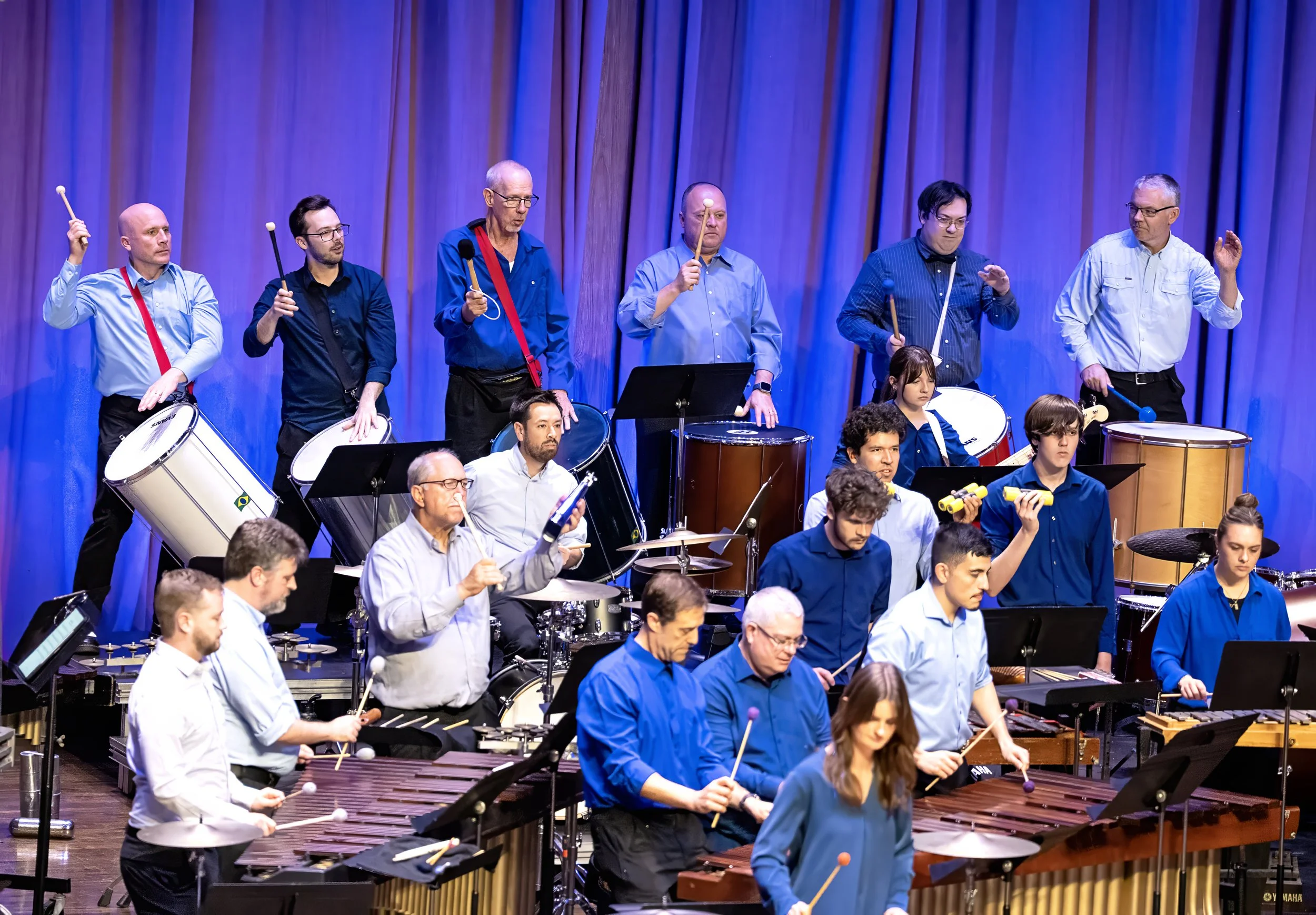 Group of musicians performing on stage with a blue curtain backdrop, playing percussion and string instruments.