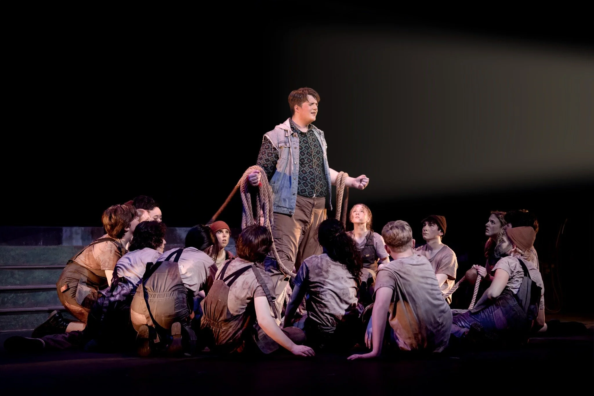 A young person stands on stage surrounded by others, holding a rope, with a spotlight shining on them.