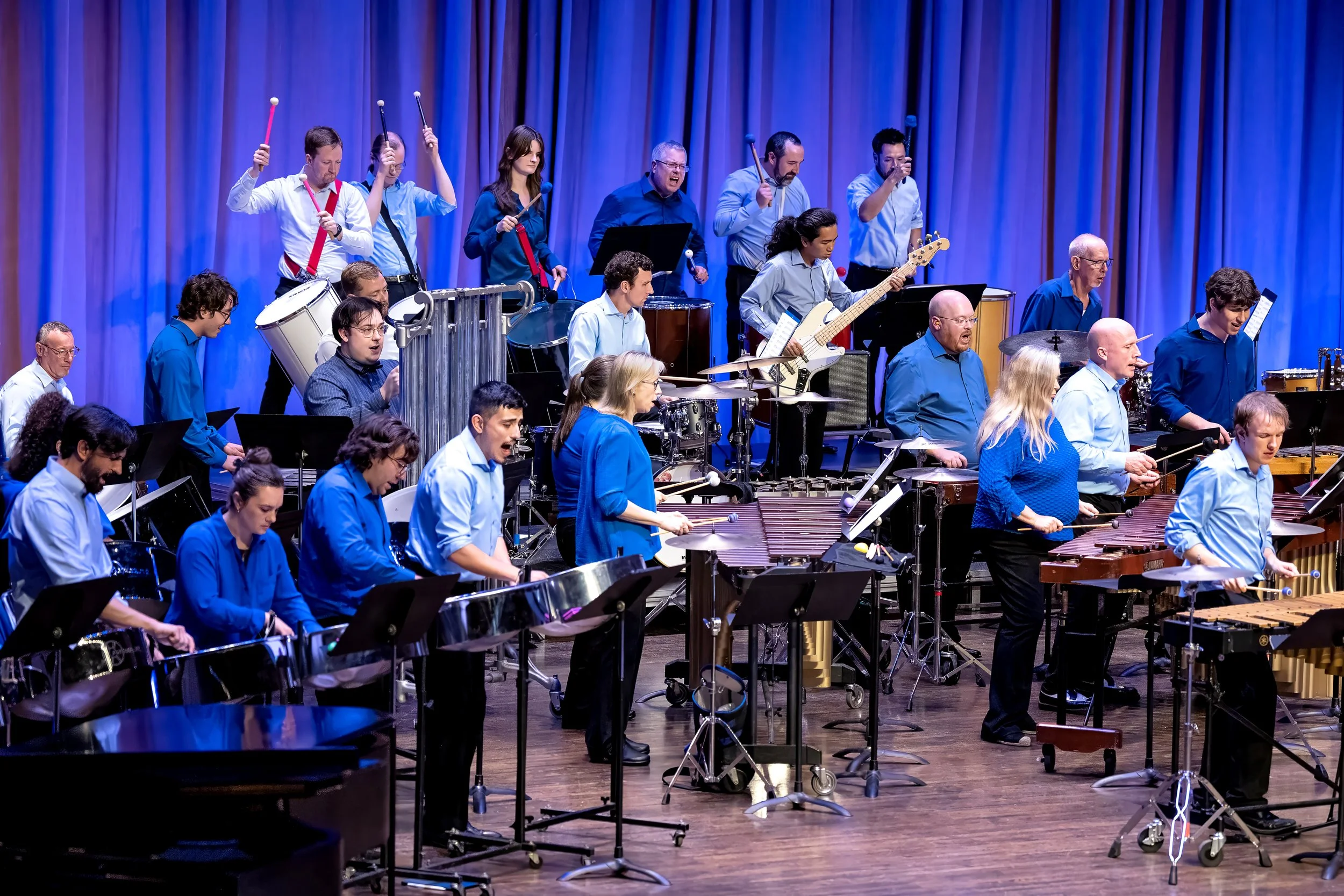 A large orchestra of musicians performing on stage with blue curtains in the background. Musicians are playing various instruments including drums, bass guitar, marimbas, and xylophones. The performers are dressed in blue or white shirts.