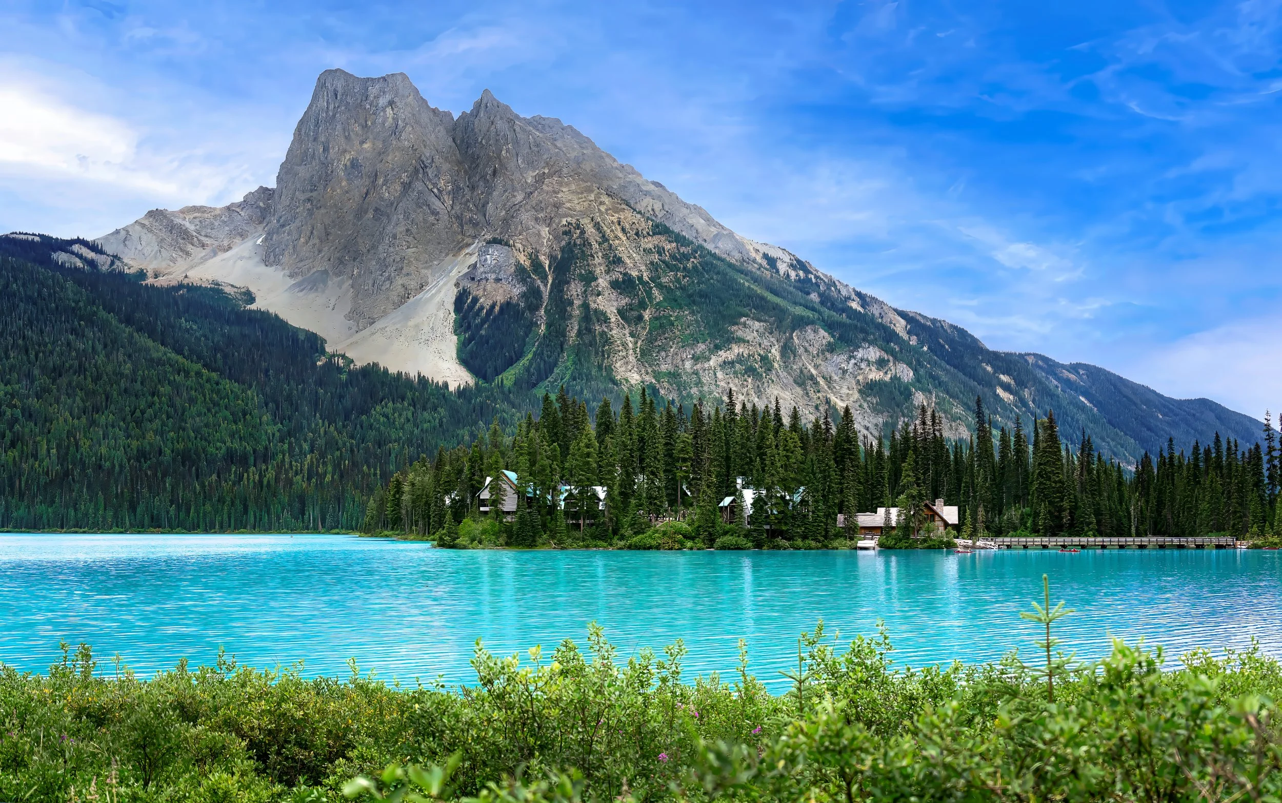 A mountain with a rocky peak over a forested area near a turquoise lake, with houses on the lake's shore and a blue sky overhead.