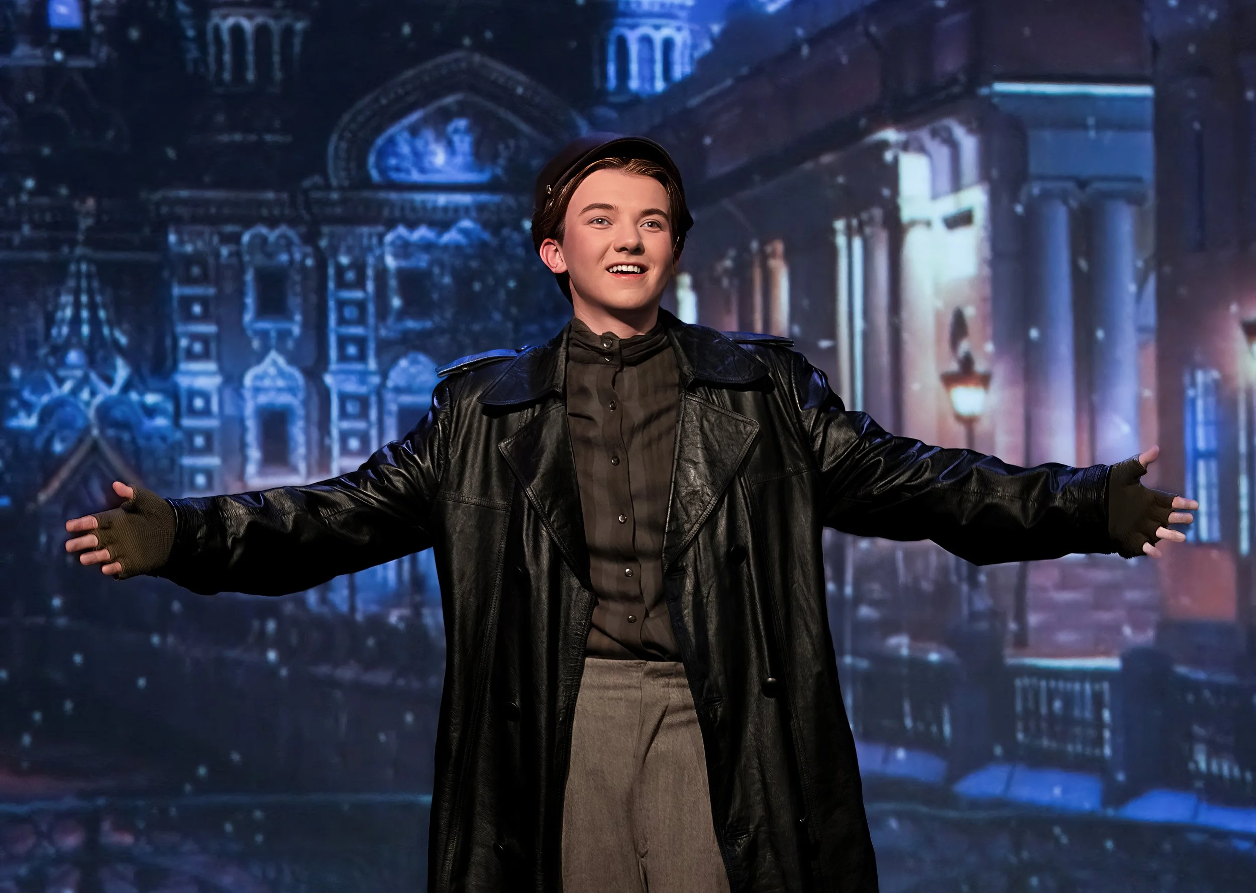 A young man with short dark hair wearing a black leather jacket, black shirt, and black headphones, standing with arms open in front of a rainy street scene at night with historic buildings illuminated by blue and warm lights.