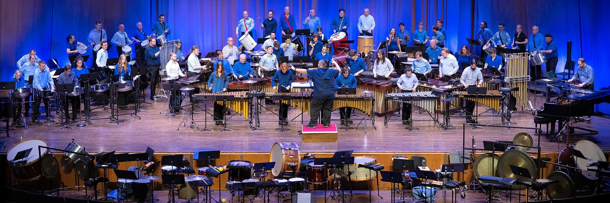 Orchestra performing on stage with conductor in front, surrounded by various percussion instruments.