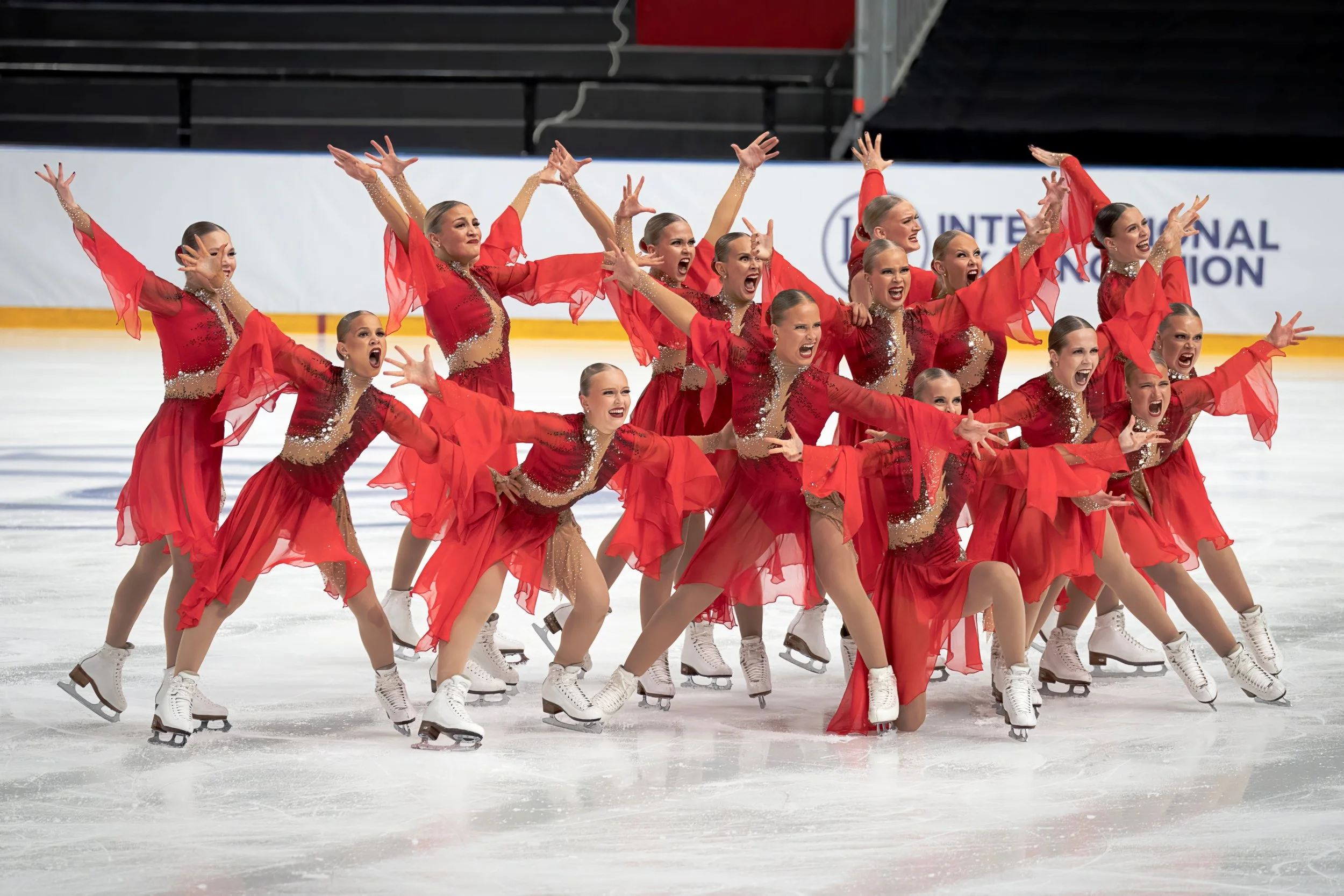 A group of female figure skaters performing a synchronized routine on ice, wearing matching red dresses with sheer long sleeves and glitter accents.