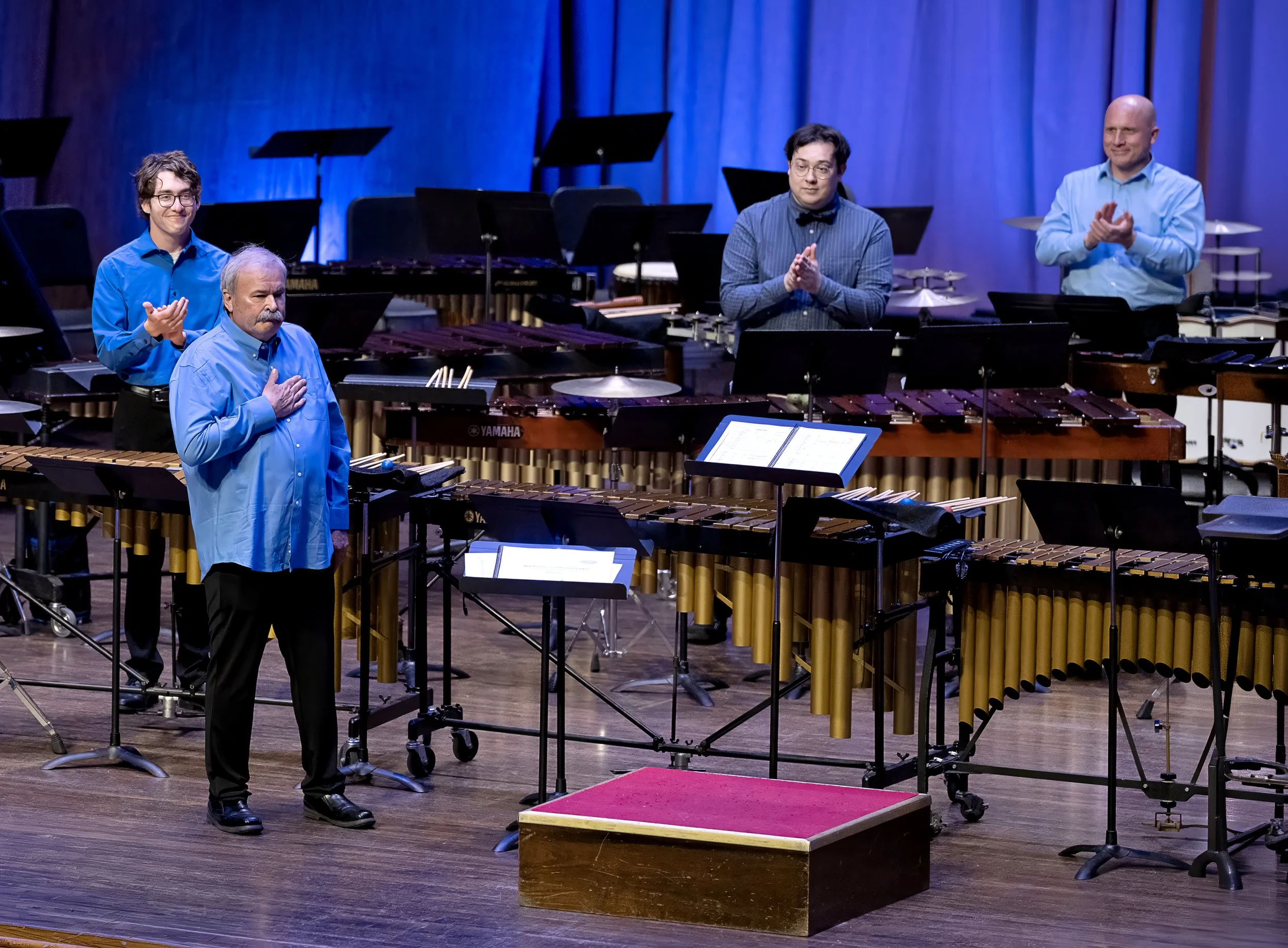 Five men dressed in blue and gray shirts standing behind percussion instruments on a stage, with a pianist and music stands in the background.
