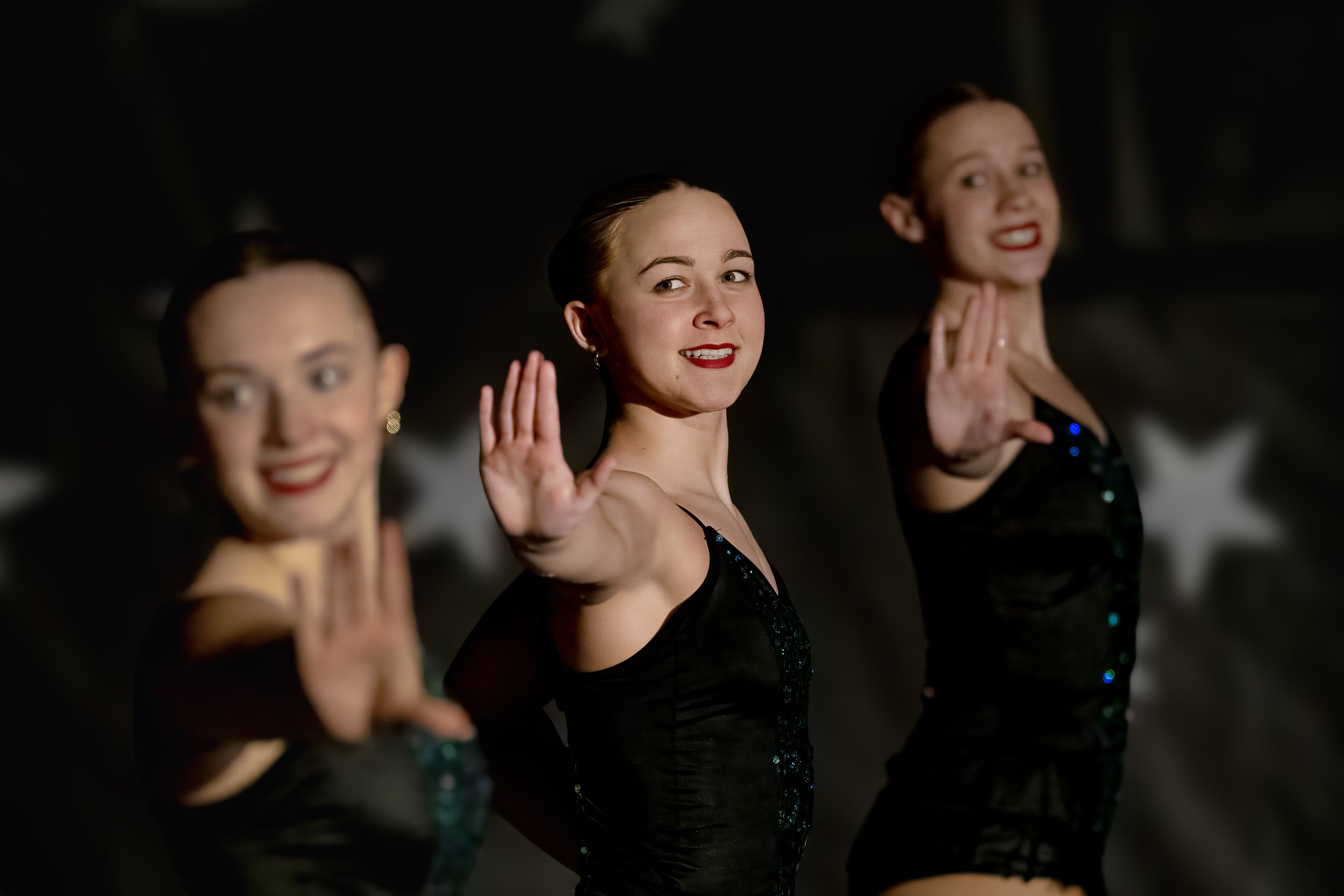 Three women in black dresses standing in a line, waving at the camera with smiles on their faces, during a performance or celebration in a dark setting.