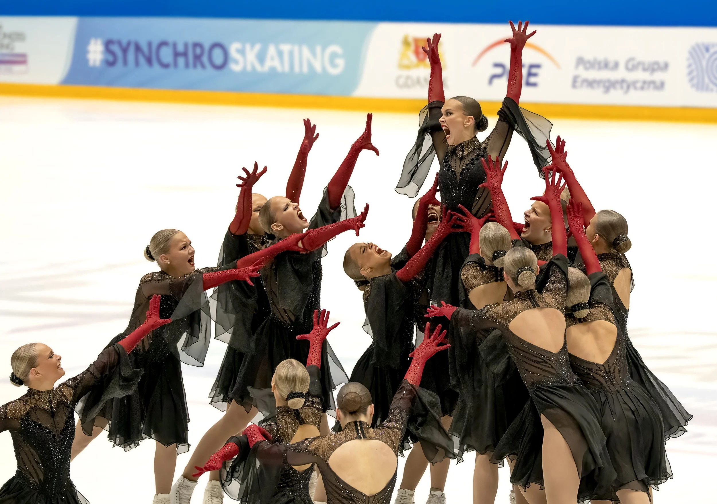 Figure skaters celebrating during a performance with a banner in the background that says '#SYNCHROSKATING'.