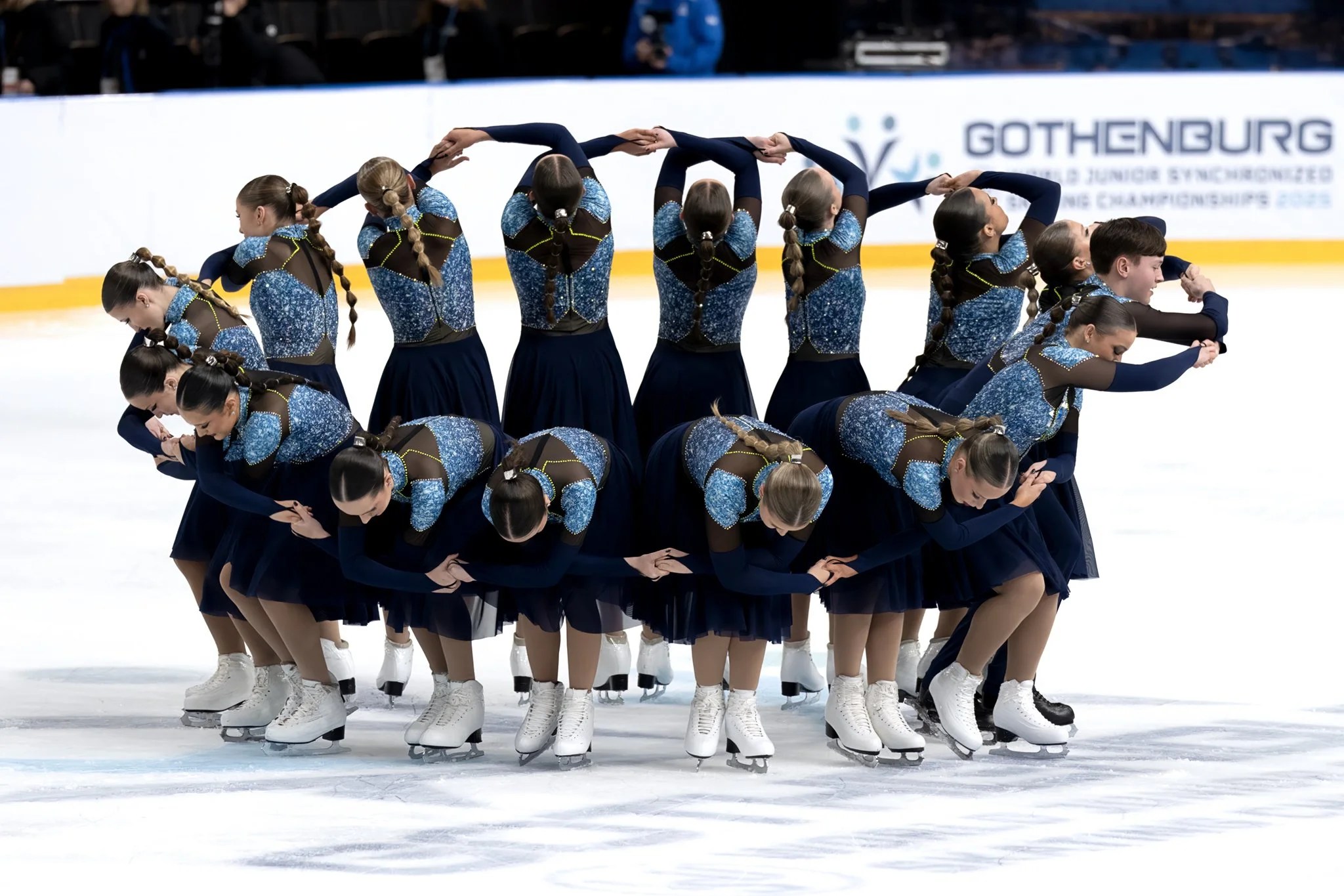 A group of young female figure skaters in matching blue and black costumes performing a synchronized routine on ice.