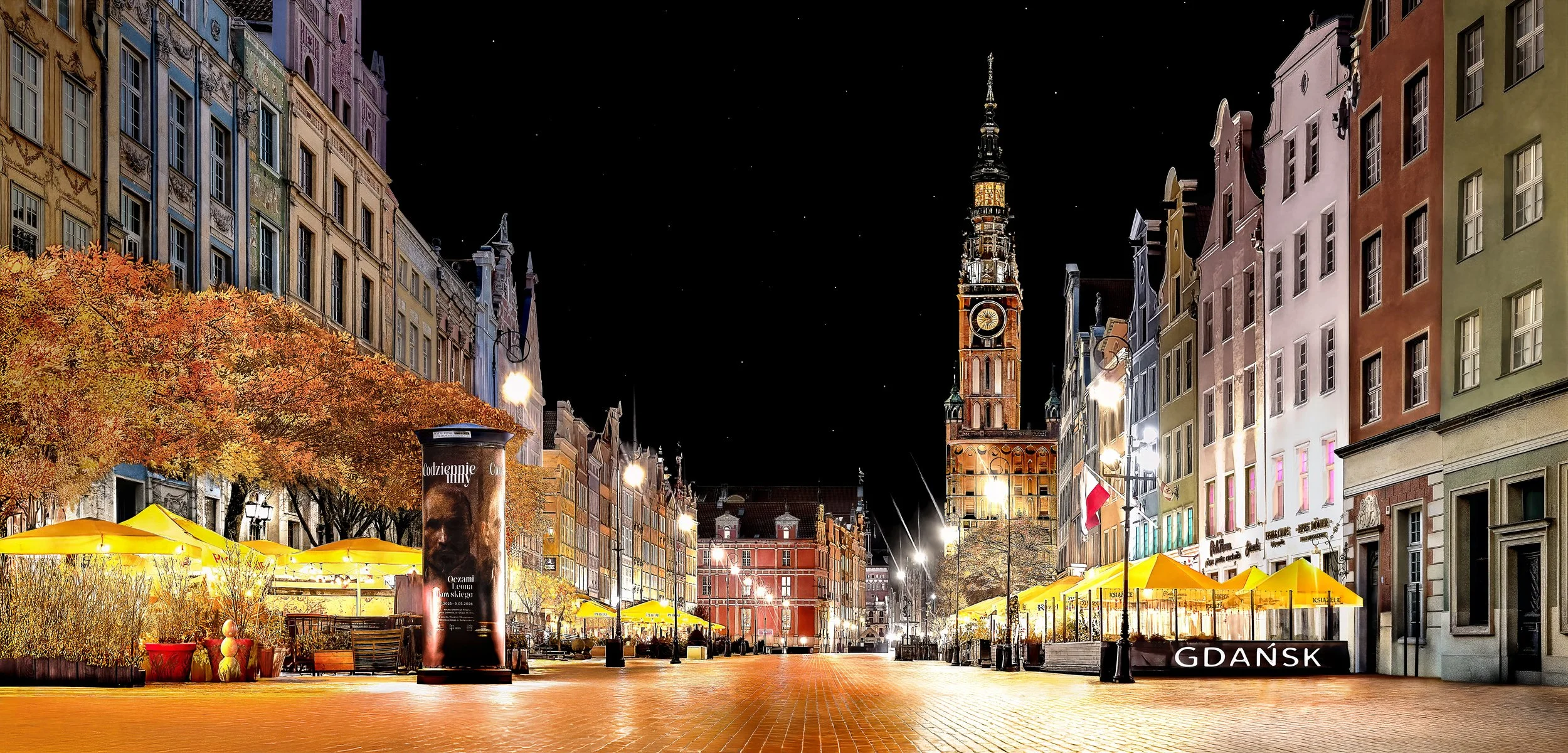 Night scene of a historic city square in Gdańsk, Poland, with colorful buildings, a tall church tower, yellow umbrellas at an outdoor market, and street lamps illuminating the area.