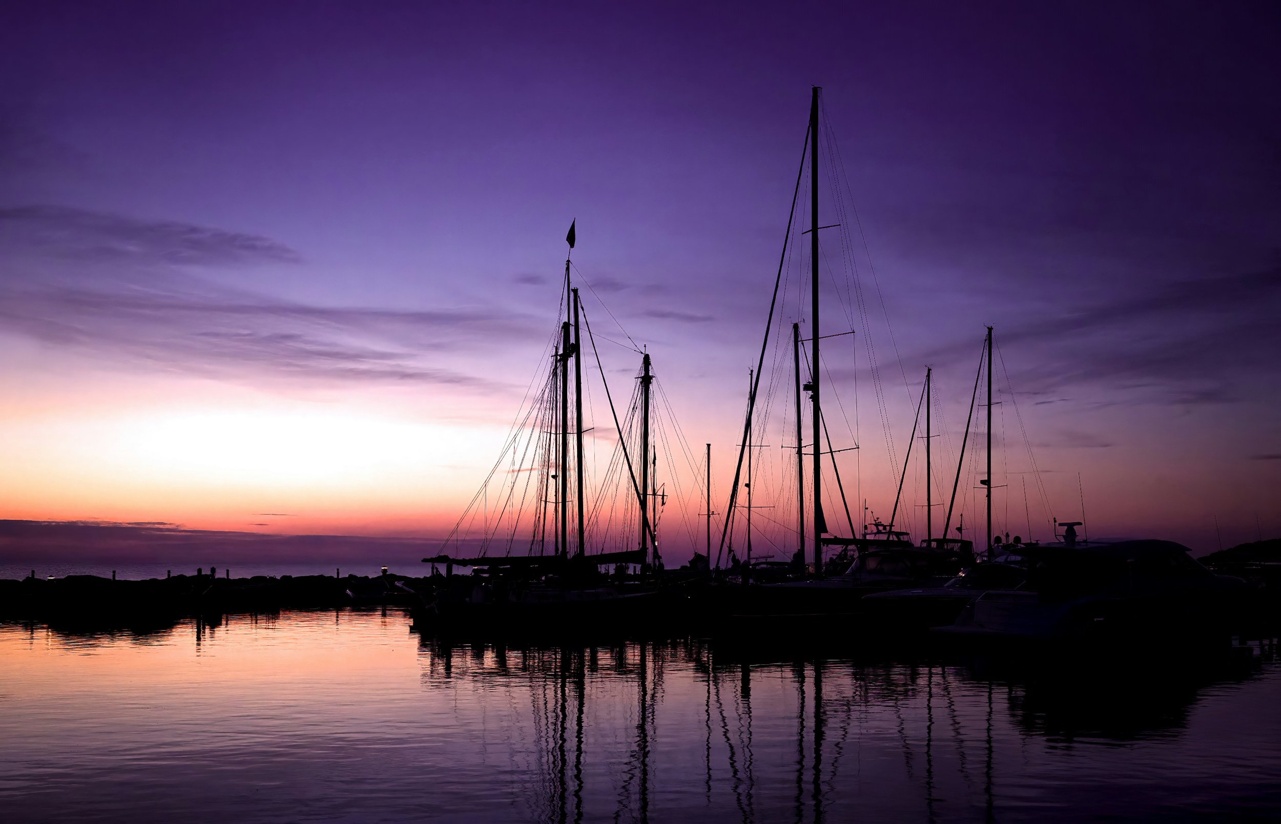 Silhouette of sailboats anchored at a marina during sunset with the sky painted in shades of purple, pink, and orange, reflected in the calm water.