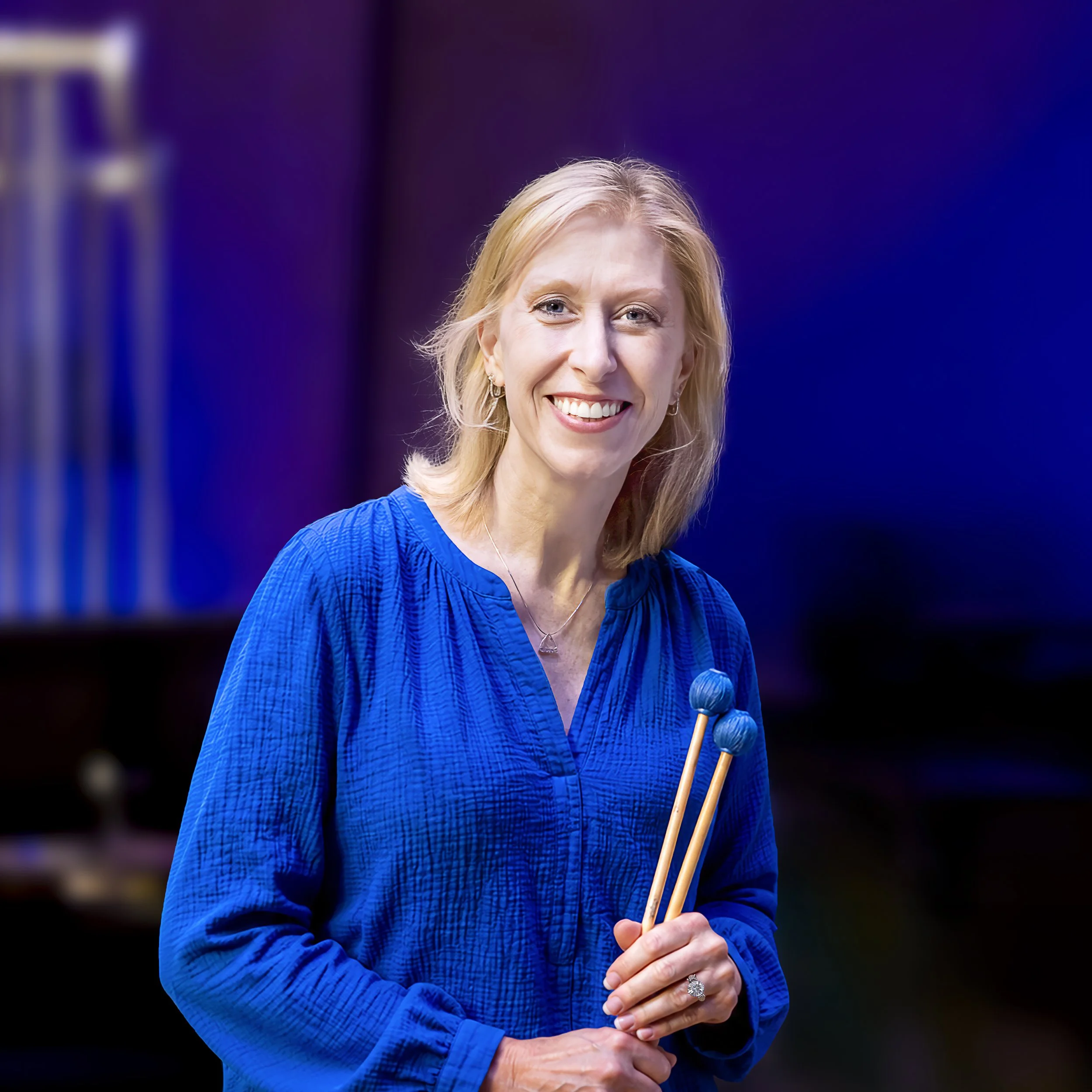 A woman with blonde hair smiling, wearing a blue patterned jacket, holding a small photo or card and a wooden stick with a decorative round top, with a blurred colorful background.