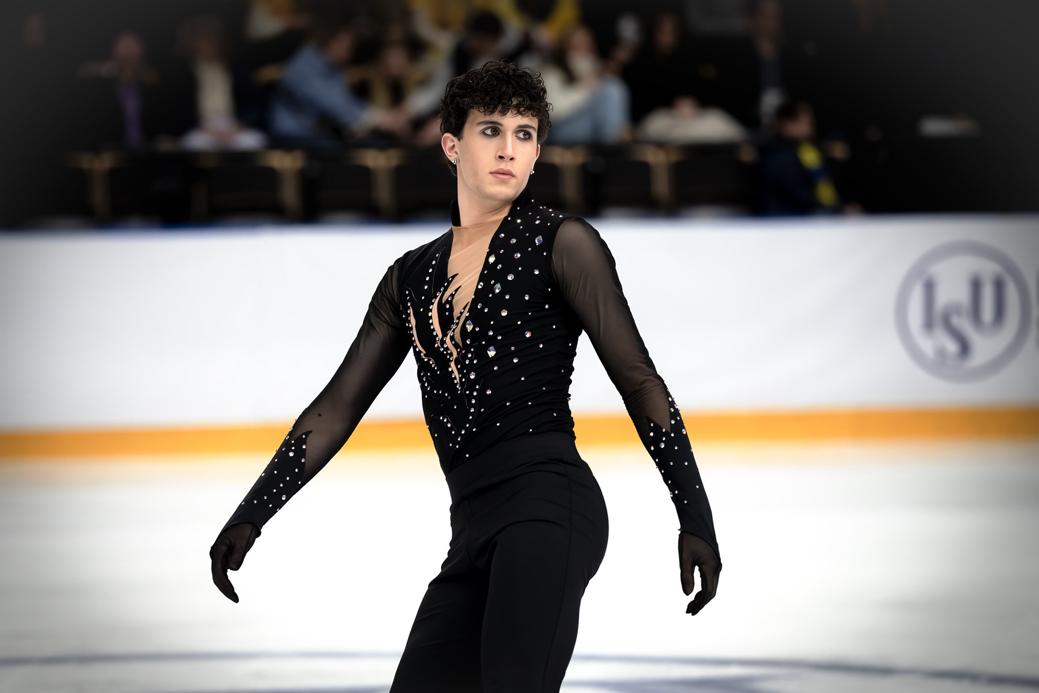 A female figure skater performing on ice rink, wearing a black, rhinestone-studded costume with sheer sleeves.