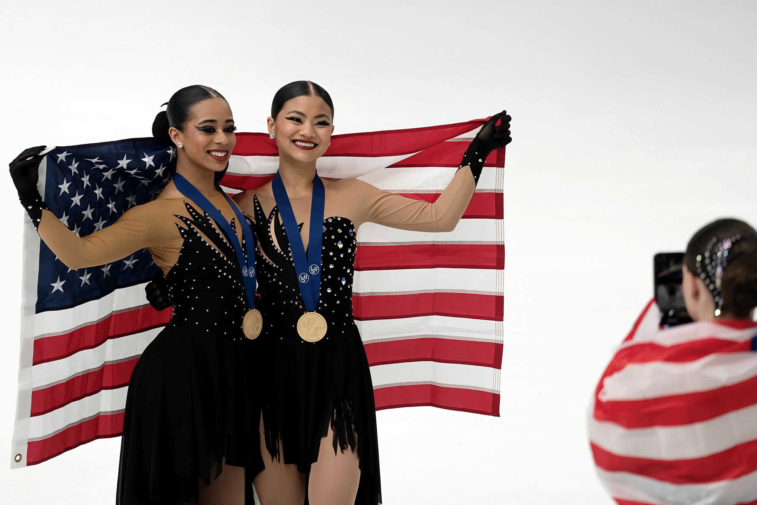 Two female figure skaters with medals around their necks pose with an American flag in the background. One skater has dark hair in a bun and the other has dark hair in a ponytail. They smile at a photographer.