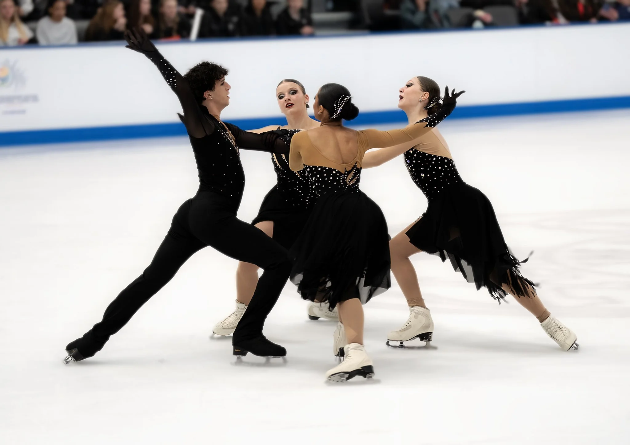 A synchronized ice skating performance featuring four female skaters in matching costumes, performing on ice with fans watching in the background.