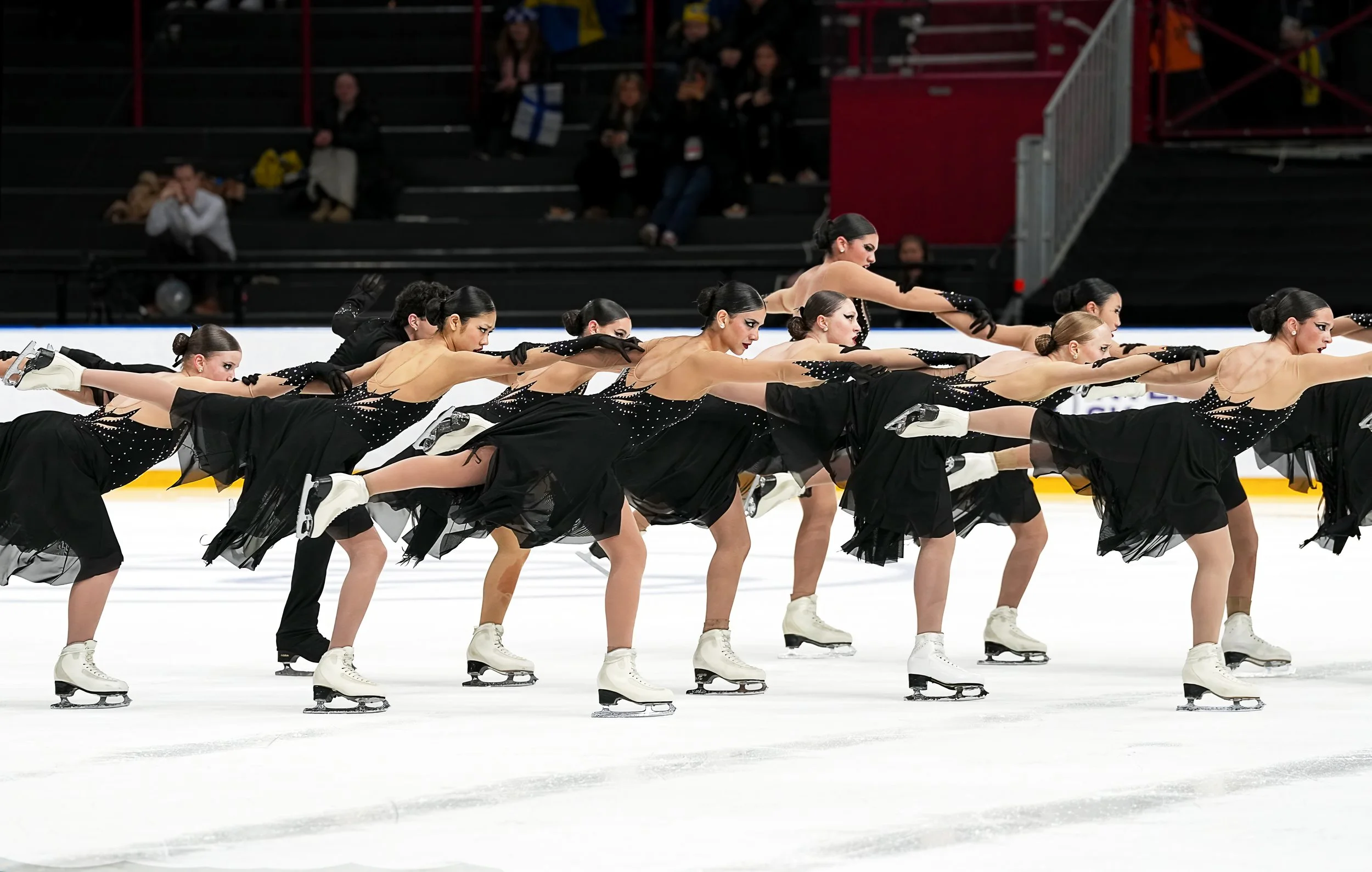 A synchronized figure skating team performing on ice rink with all members in similar pose, wearing matching black and beige costumes with sparkles, black gloves, and skating boots.
