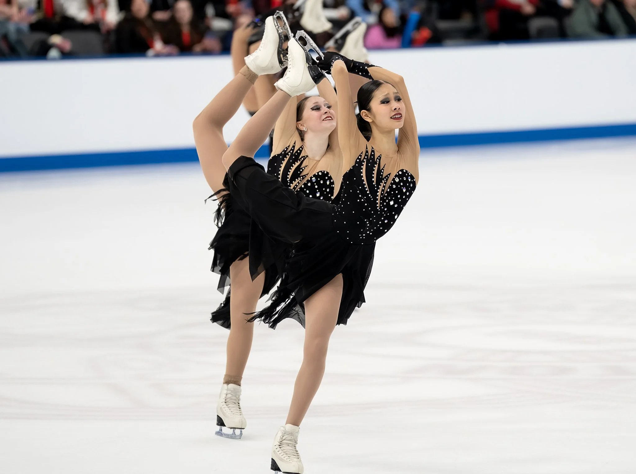 Two female figure skaters performing a lift during a routine on ice rink, both wearing black costumes with silver embellishments, one with hair tied back and the other with hair down, and both wearing white figure skates.