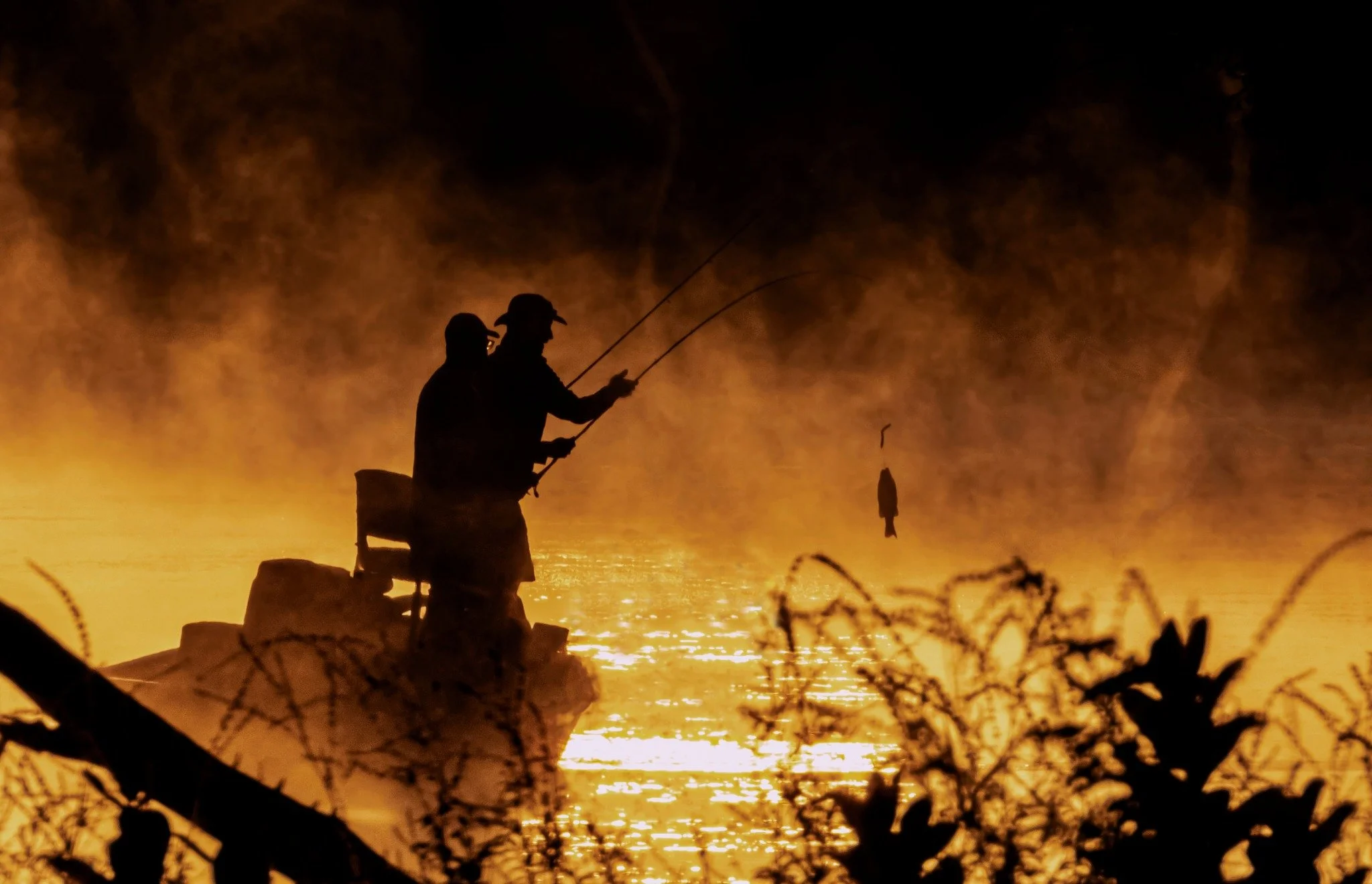 Two people fishing at sunset on a body of water, silhouetted against an orange sky with reflections on the water surface.