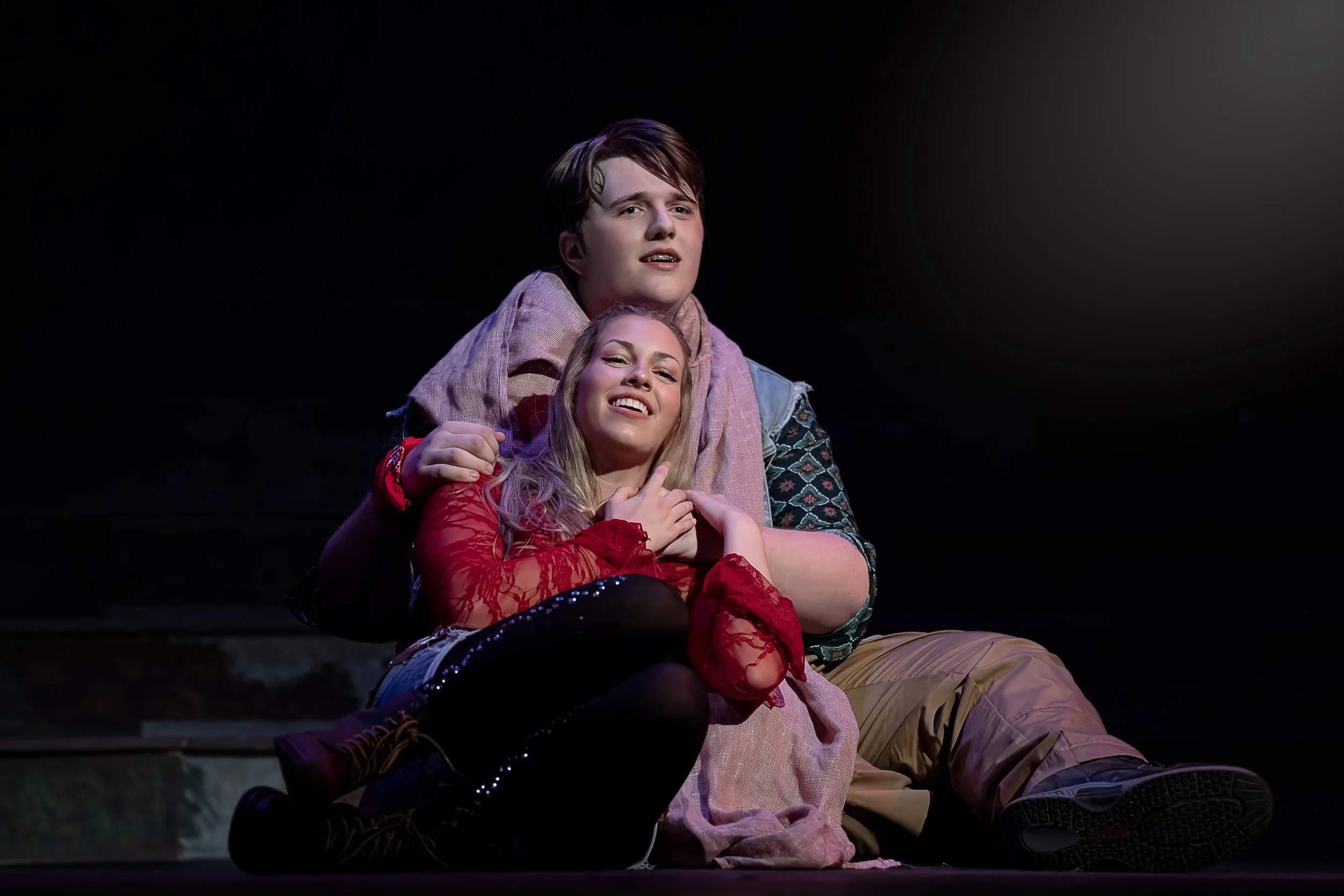 Two young actors on stage, one male and one female, with emotional expressions. The male has short hair and the female has long, blonde hair, both dressed in colorful costumes. The background is dark.