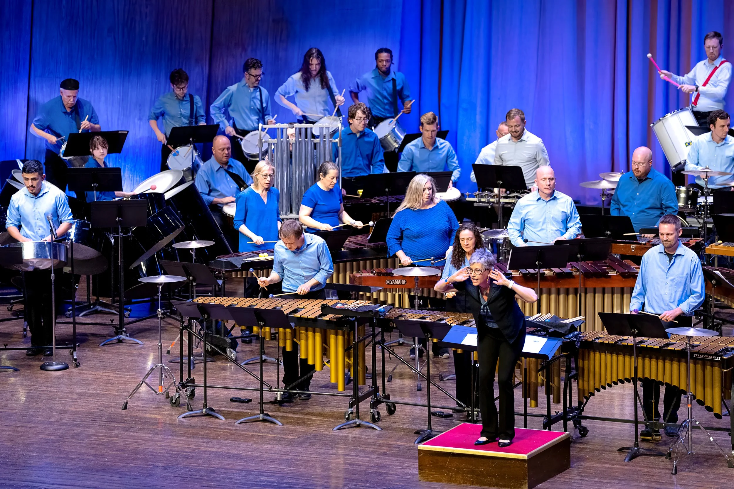 An orchestra performing on stage with musicians playing percussion instruments, led by a conductor standing on a red podium.