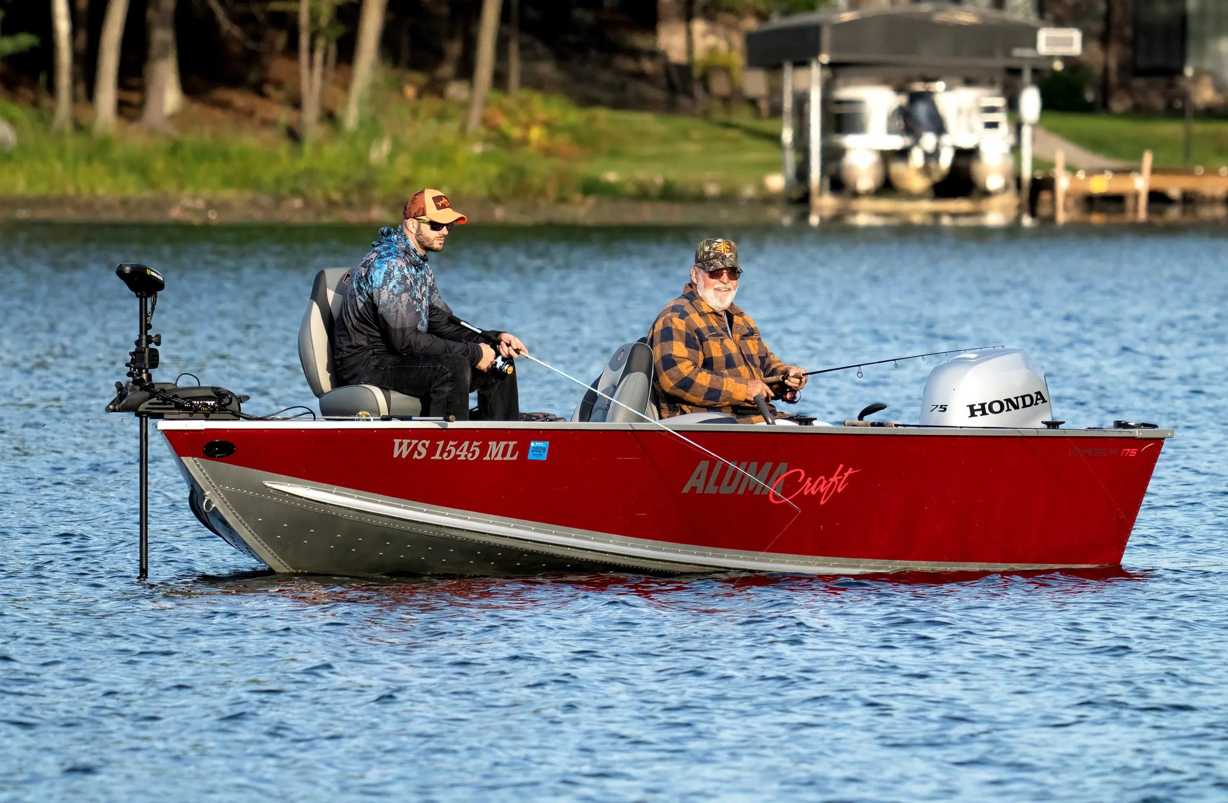 Two men fishing on a red boat with a Honda outboard motor on a lake, with trees and a boathouse in the background.