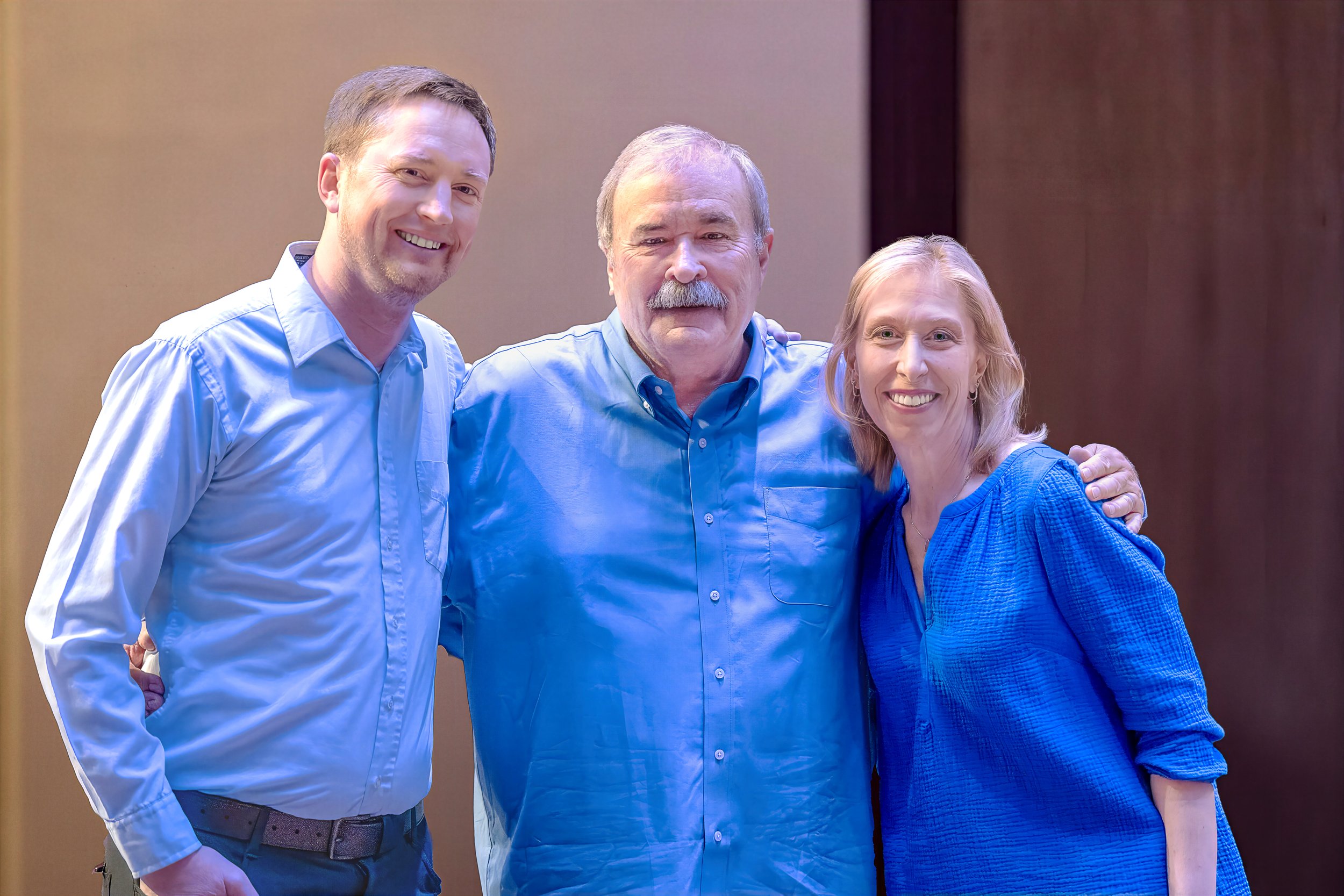 Three people standing together smiling, two men and one woman, all wearing blue shirts, in an indoor setting.