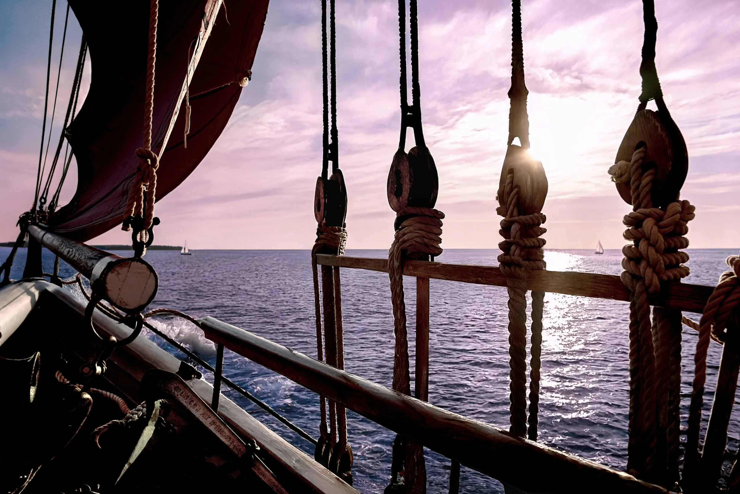View of the ocean from a sailing ship with ropes and pulleys in the foreground, the sky has pink and purple clouds with the sun setting, and a small sailboat is visible in the distance.