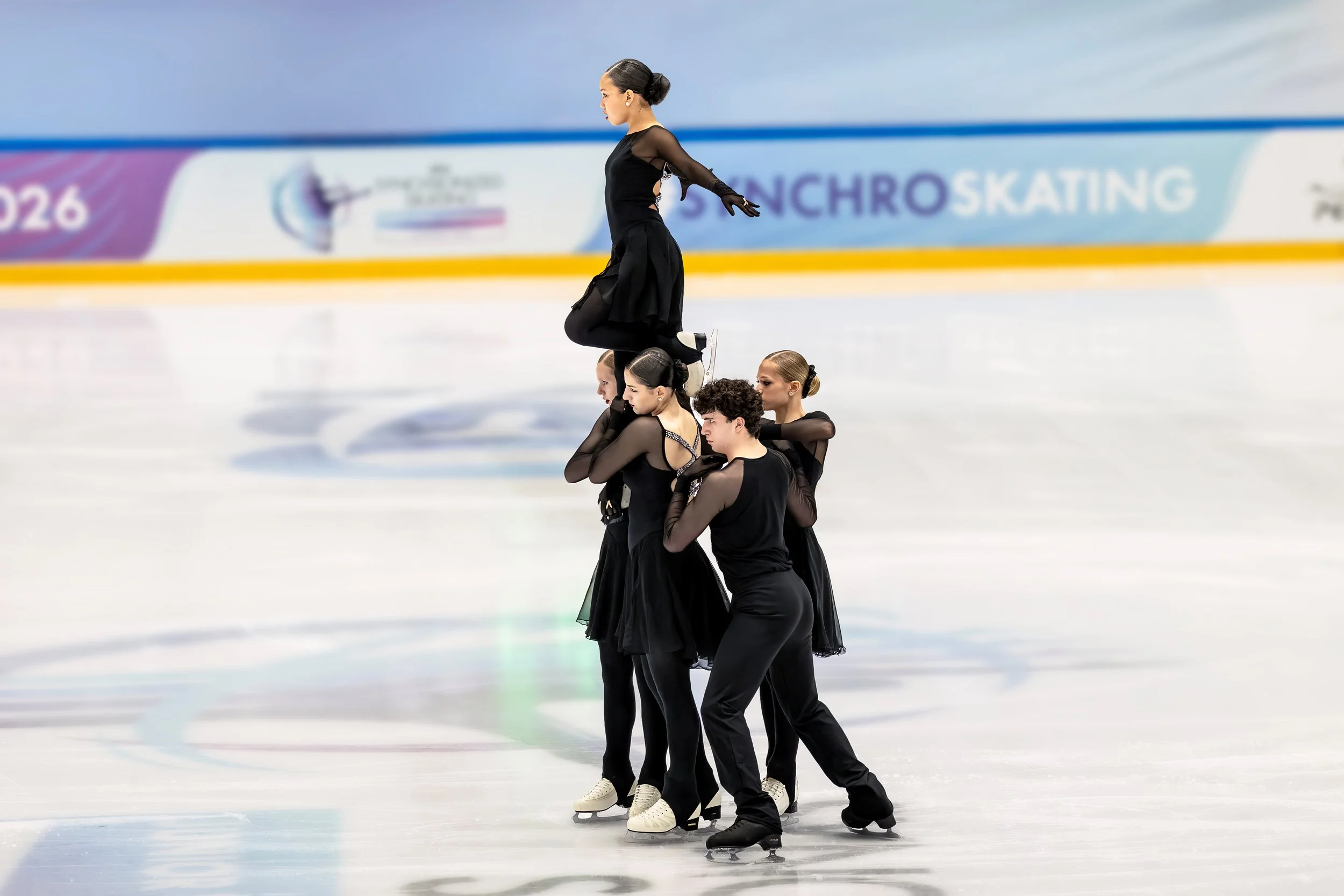A group of five young women in black outfits performing an ice skating routine on an indoor ice rink, with one woman balanced atop the others in a choreographed formation.