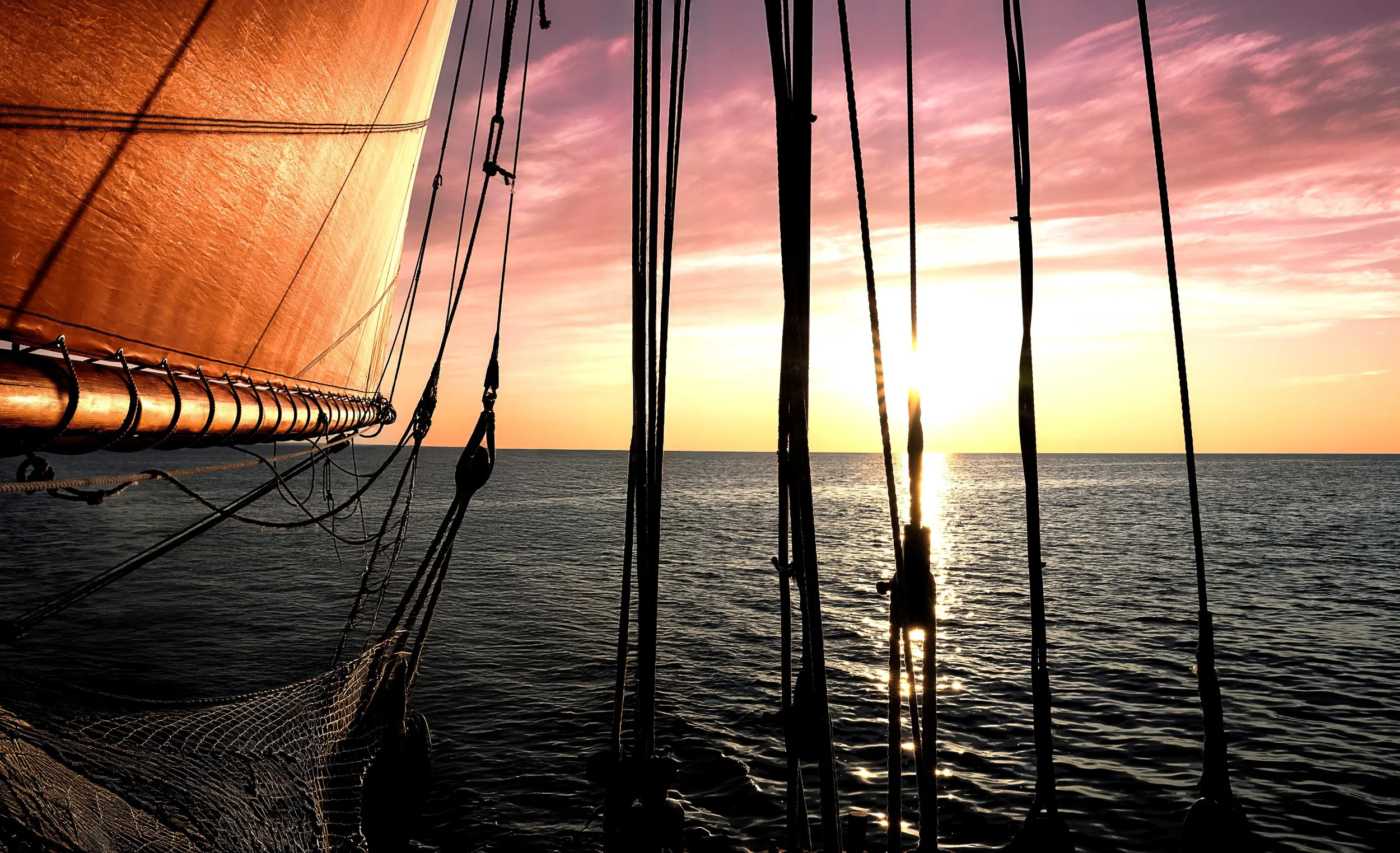 View from a sailboat at sunset with the orange sail on the left and the sunset over the water in the background, viewed through the boat's rigging and mast.