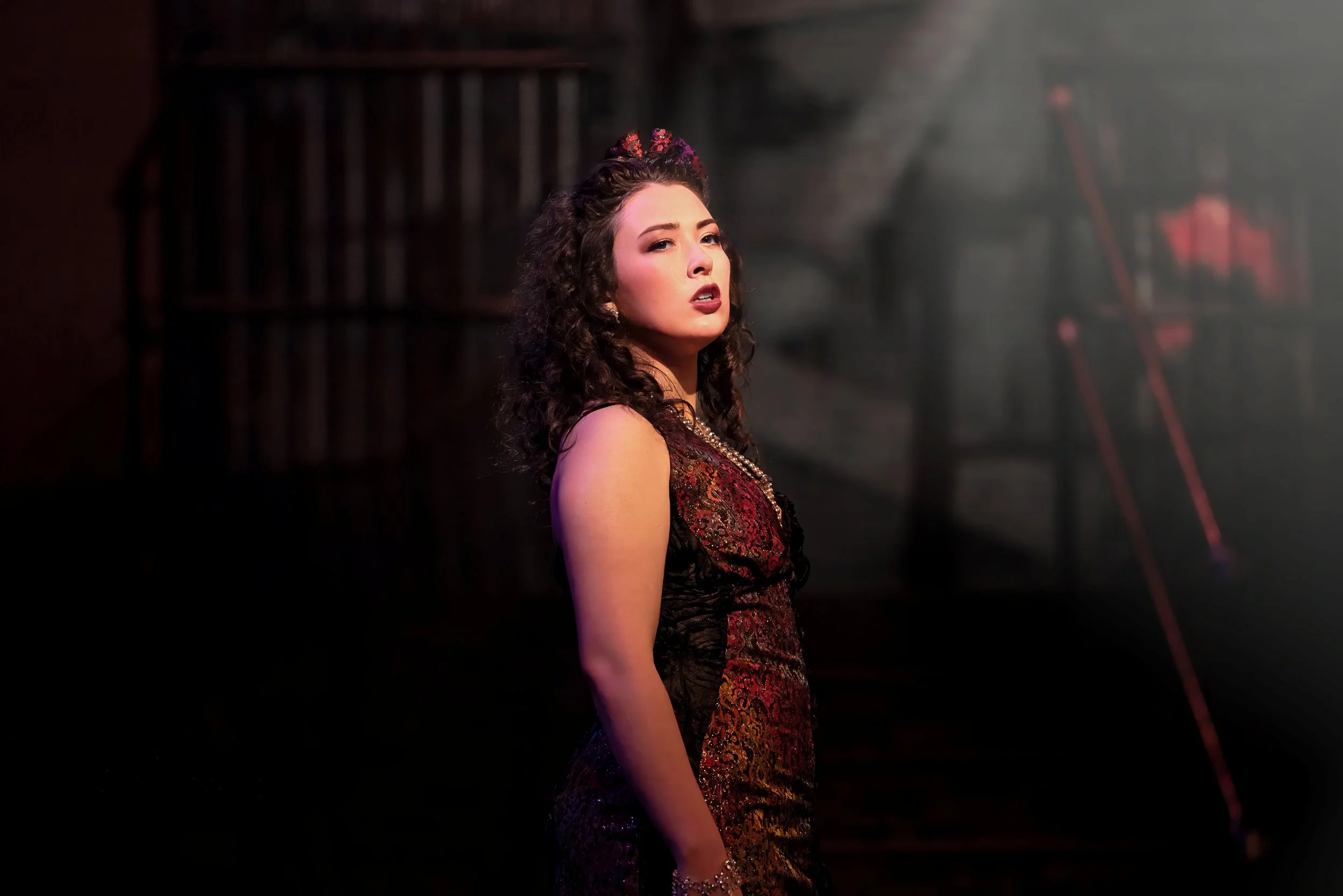 A woman with curly hair styled with a floral hair accessory, wearing a dark red and black dress with lace details, standing in a dimly lit room with a blurred background.