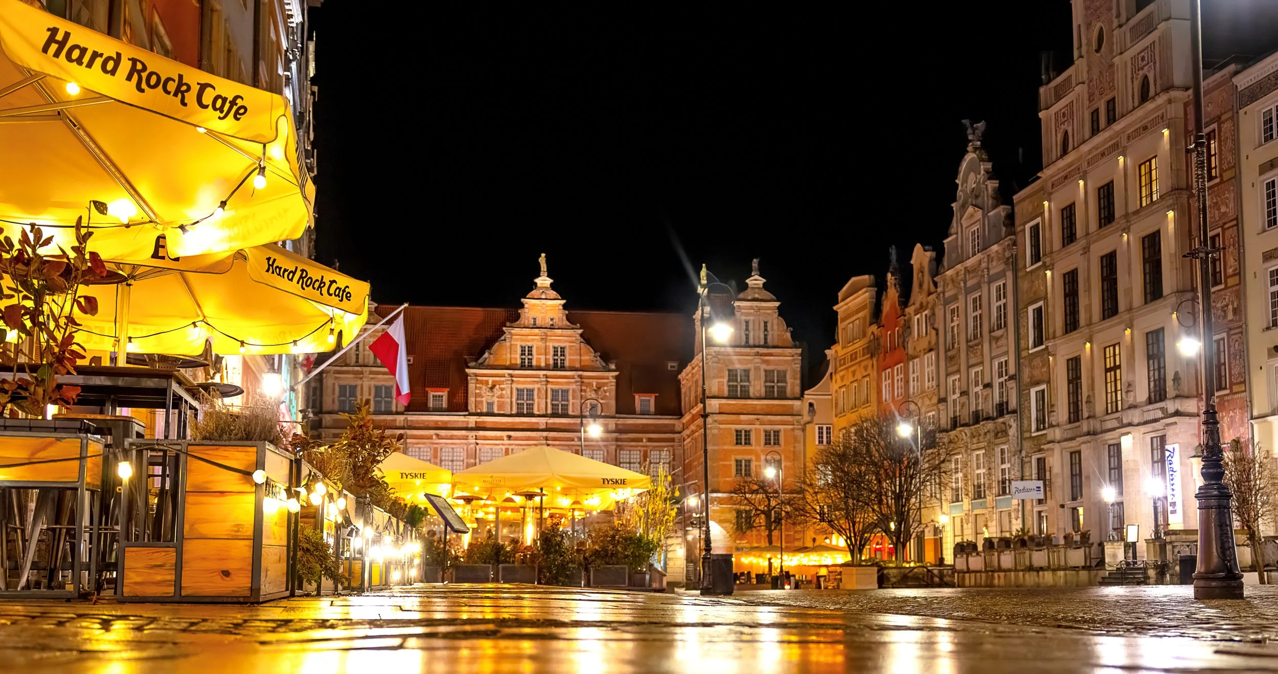 Night view of a European city square illuminated with yellow lights from outdoor cafes and street lamps, with historic buildings and a church in the background.