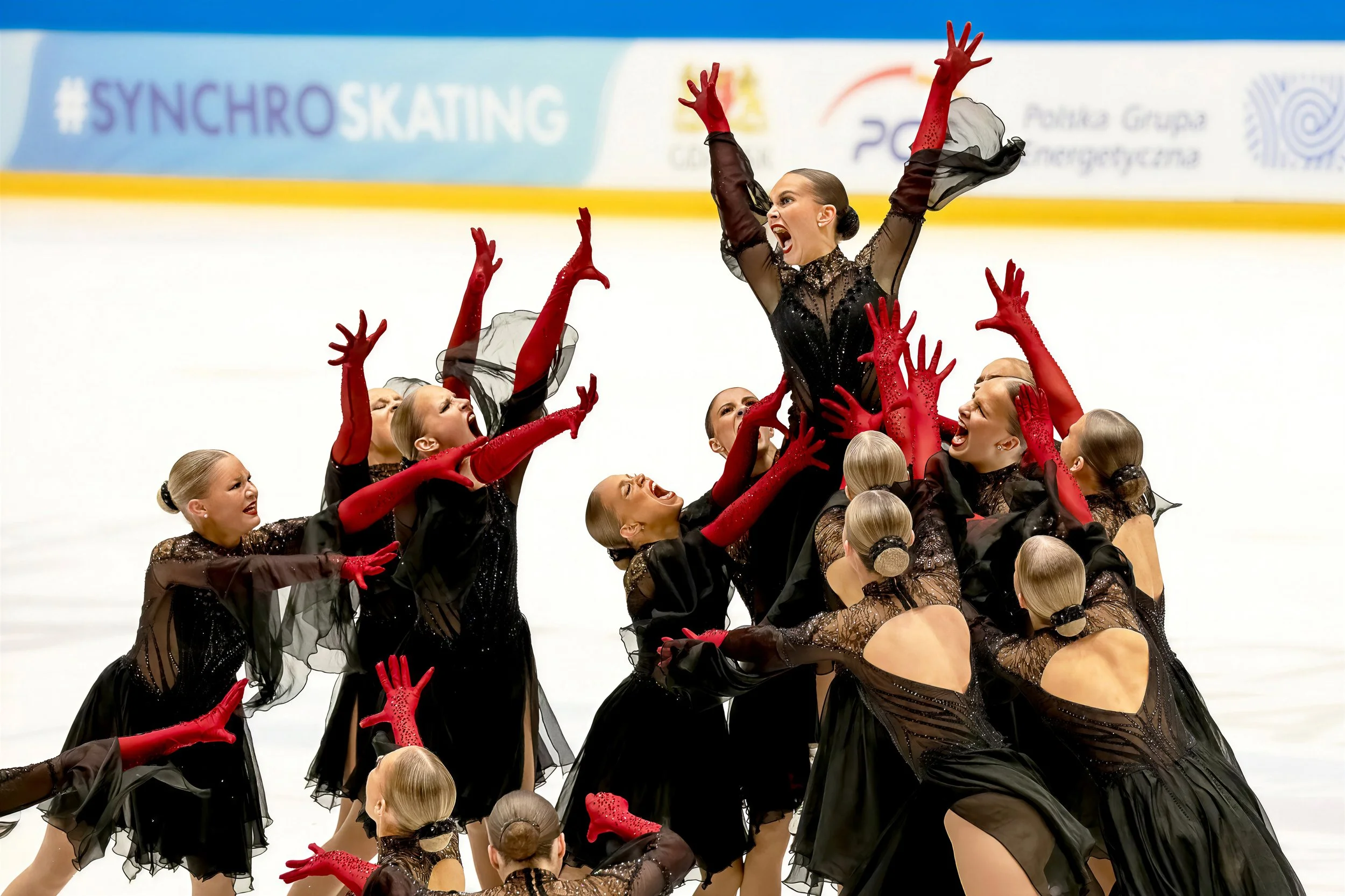 A group of figure skaters wearing black dresses and red gloves performing on ice. One skater is lifted in the air with arms outstretched, while others support her. They are all smiling and appear to be celebrating.