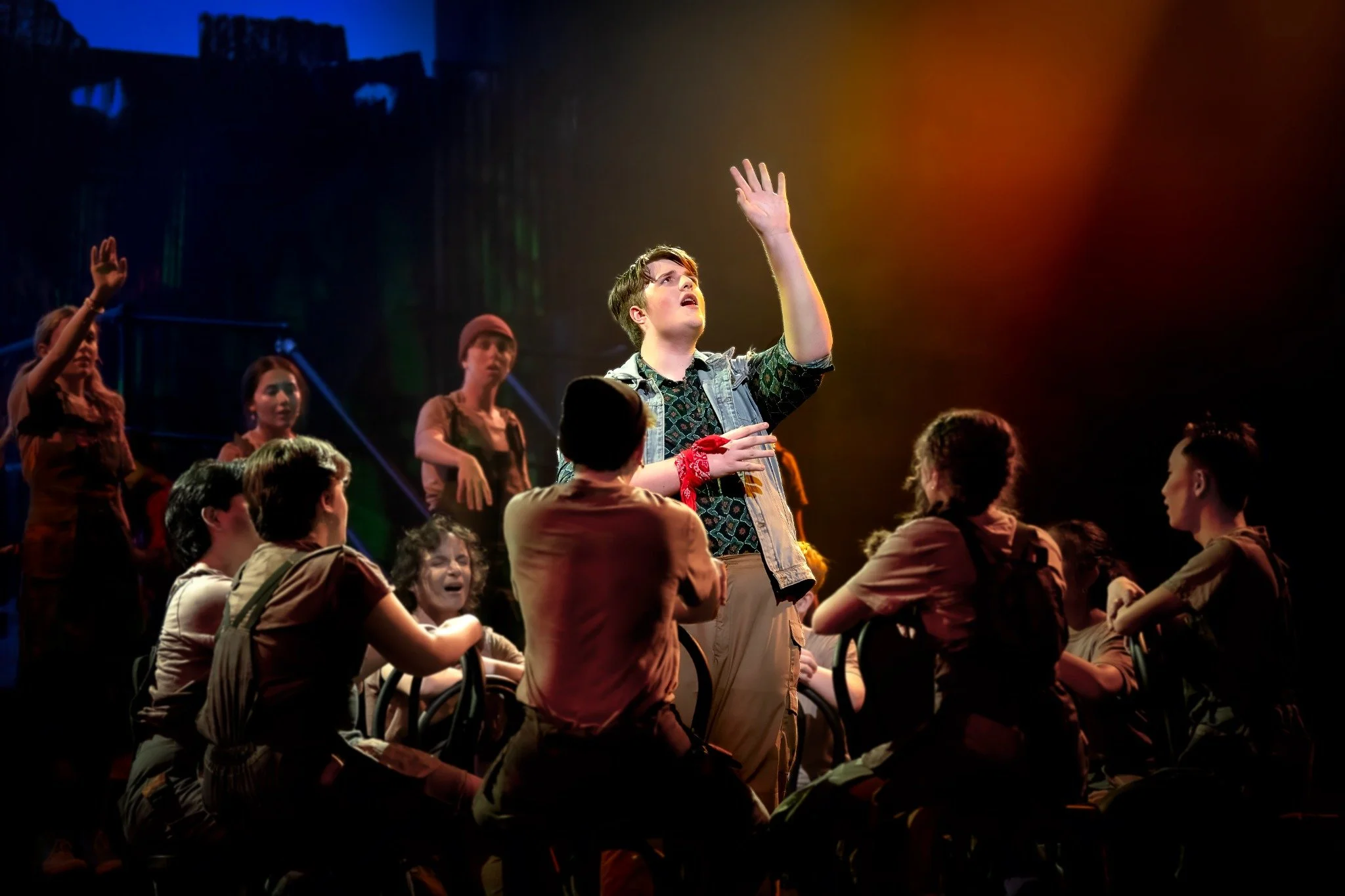 A group of young people sitting and standing in a dimly lit room with colored stage lights, some raising their hands as if in a performance or rehearsal, with a young man in the center reaching upward.