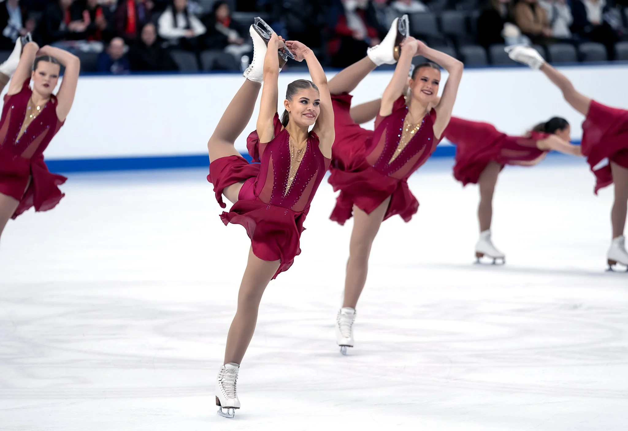 Figure skaters in matching red costumes performing a synchronized routine on ice rink with an audience in the background.