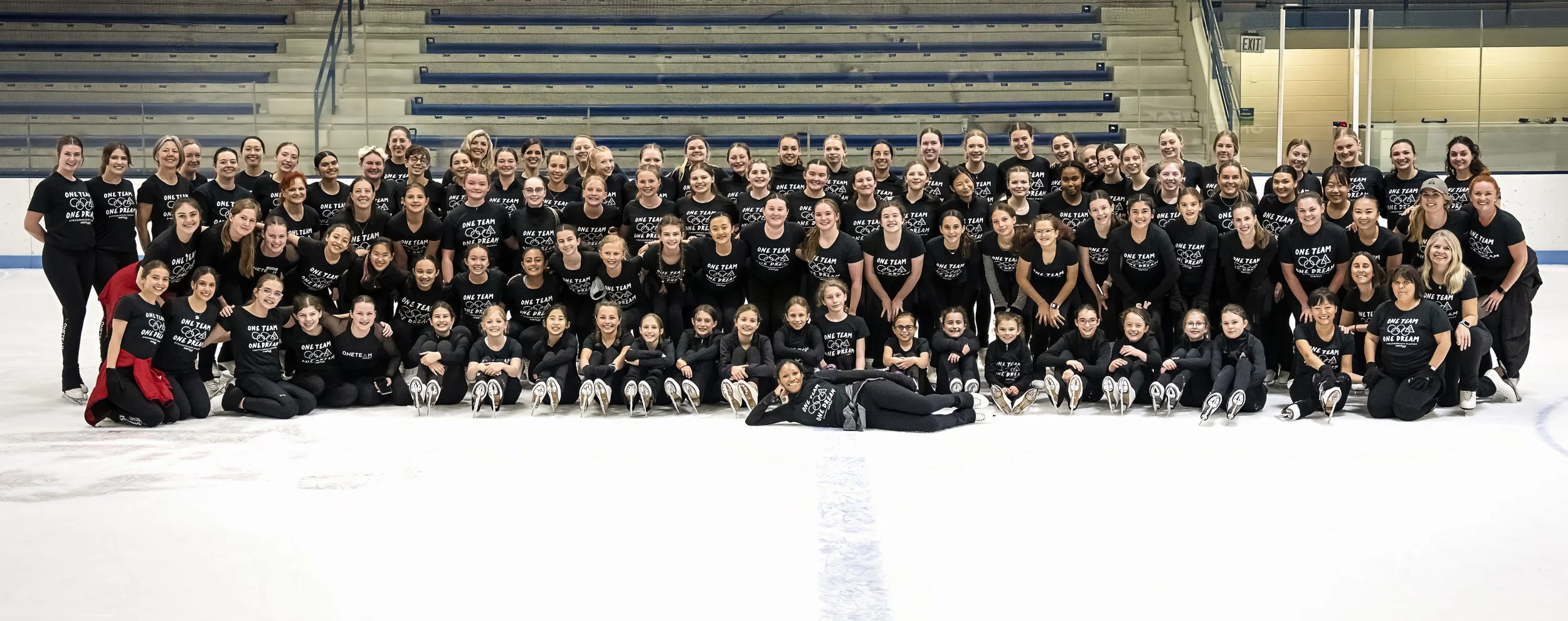 A large group of ice skaters and coaches pose together on an ice rink, wearing matching black T-shirts with a skating logo and the words "ONE TEAM ONE DREAM."