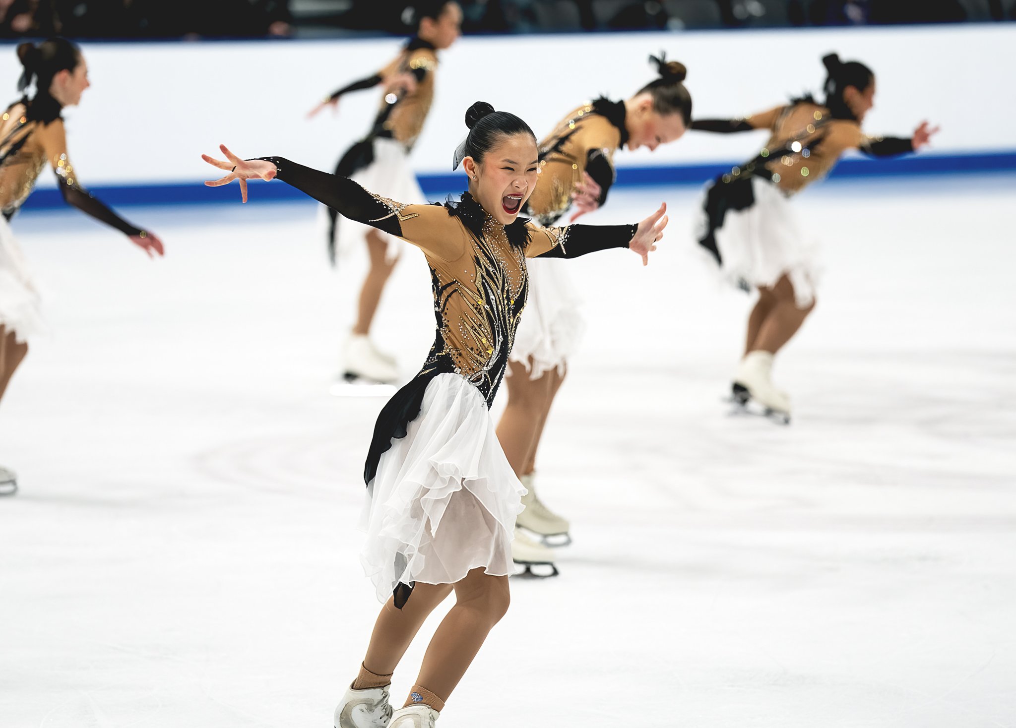 A group of female figure skaters dressed in elaborate costumes performing on the ice, with the focus on one skater in the forefront expressing intense emotion.