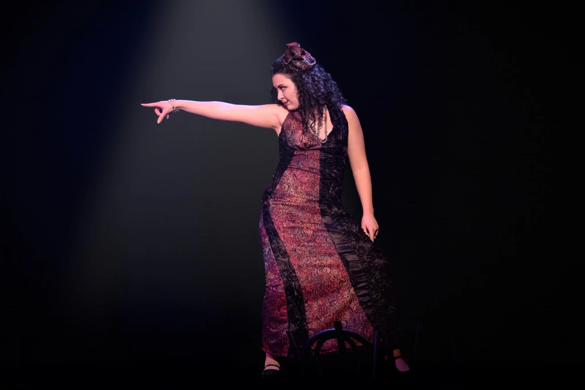A woman with curly dark hair wearing a long red and black patterned dress, standing on a dark stage with one arm extended and pointing, with a spotlight shining down on her.