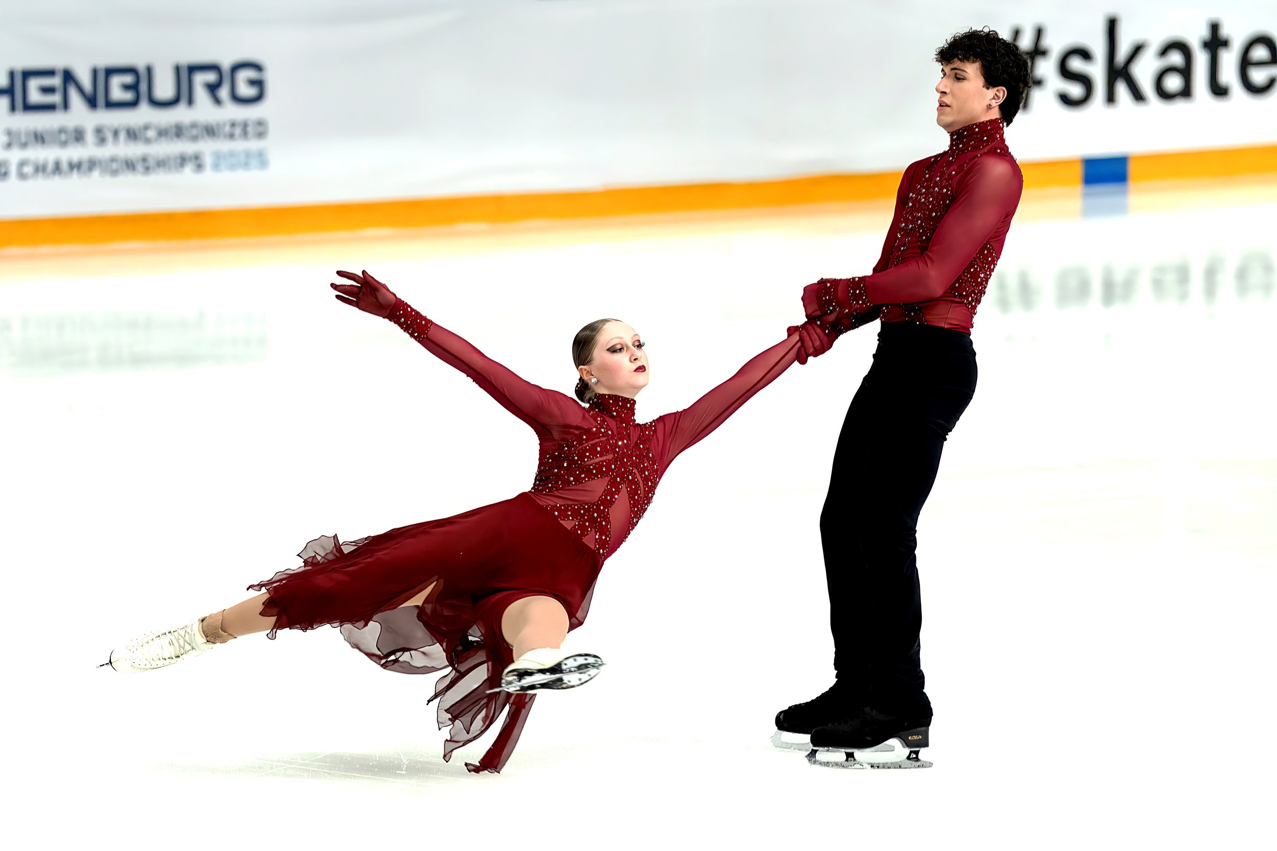 Two figure skaters, a woman in a red skating dress and a man in a black outfit, perform a dance on ice rink. The woman is in a deep lunge with arms extended, while the man holds her hands. They are skating near each other during a performance.