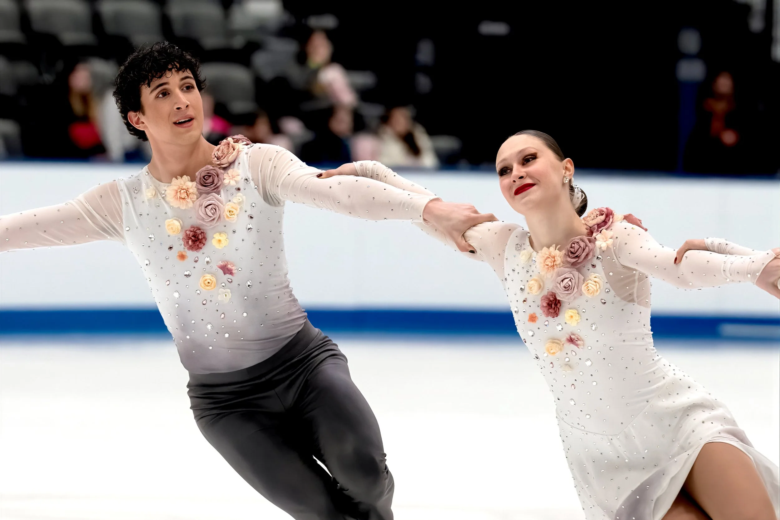 Two figure skaters, a man and a woman, performing a routine on ice. They are holding hands, wearing elegant costumes adorned with flowers and rhinestones, with the woman in a white dress with floral embellishments.