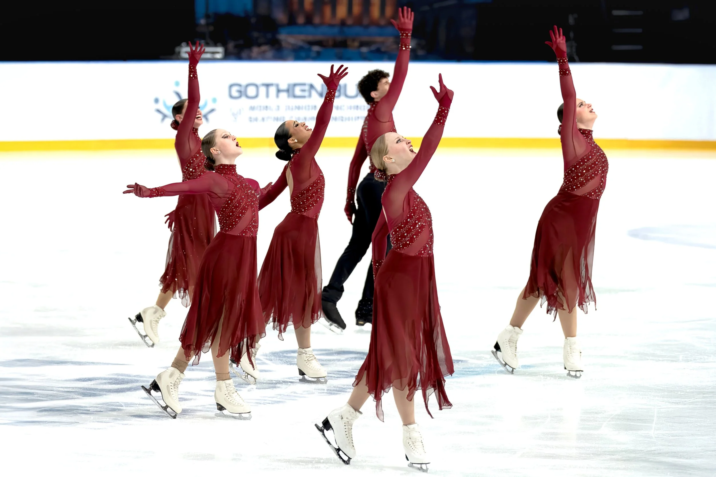 A group of female figure skaters dressed in red costumes practicing on ice rink, performing a synchronized routine with their arms raised.