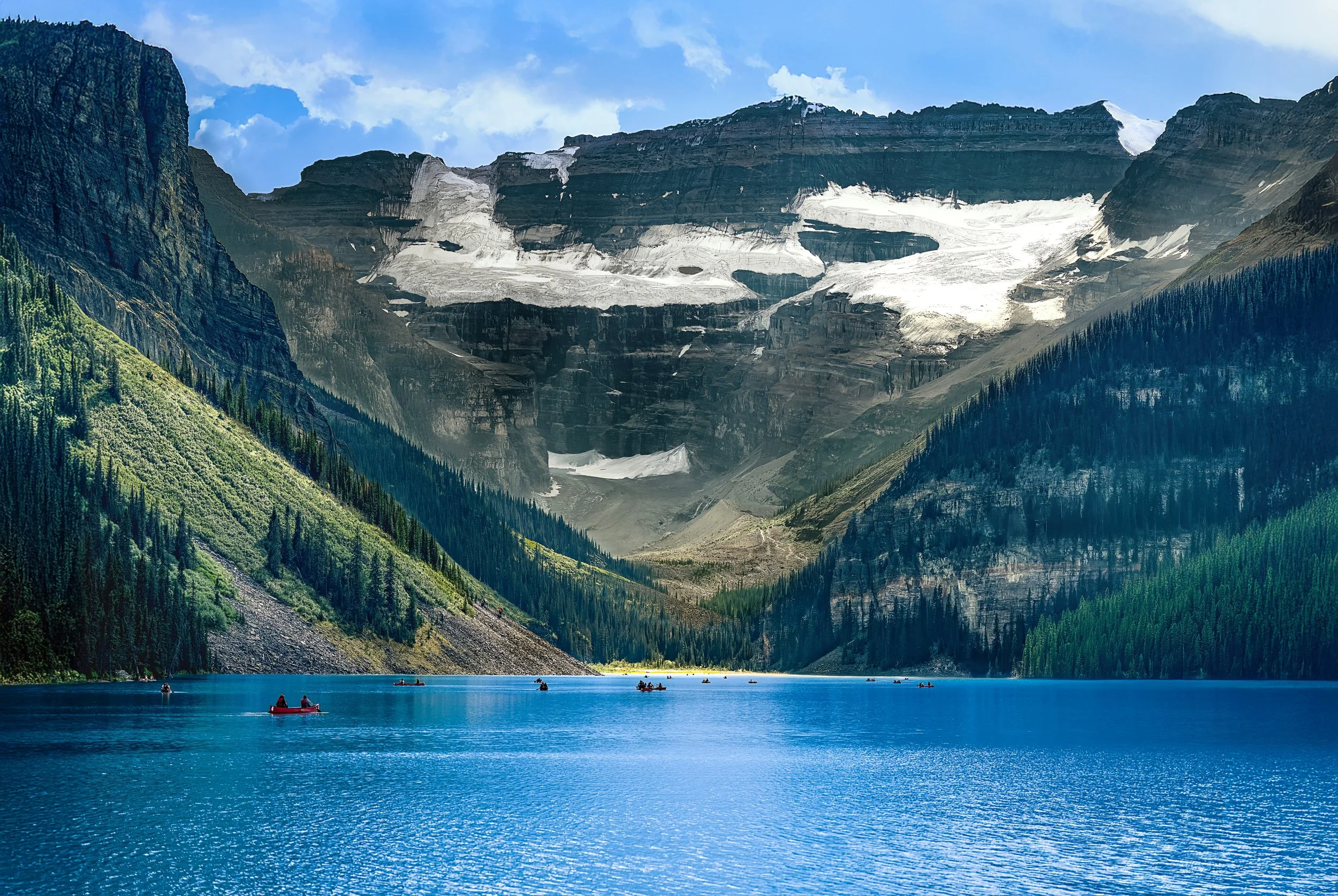 Scenic mountain landscape with glacier-covered peaks, green forested slopes, and a calm lake with small boats and people paddling.