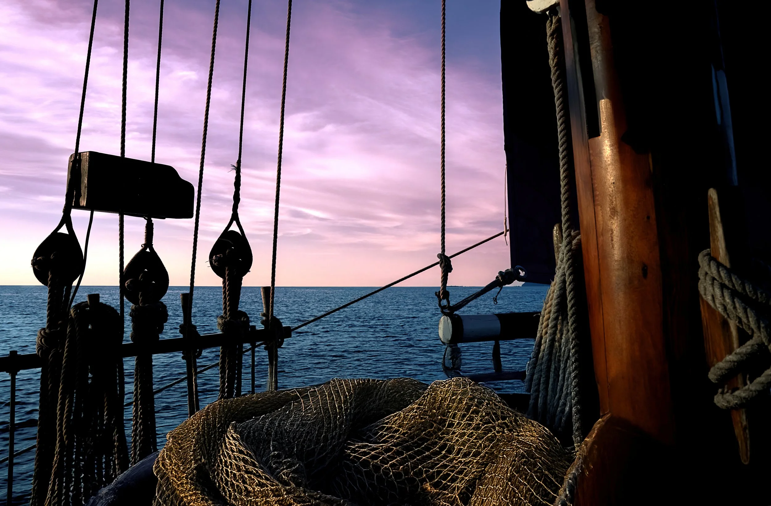View from a sailboat showing ropes, pulleys, and part of the wooden ship structure with a calm ocean and cloudy sky in the background during sunset or sunrise.