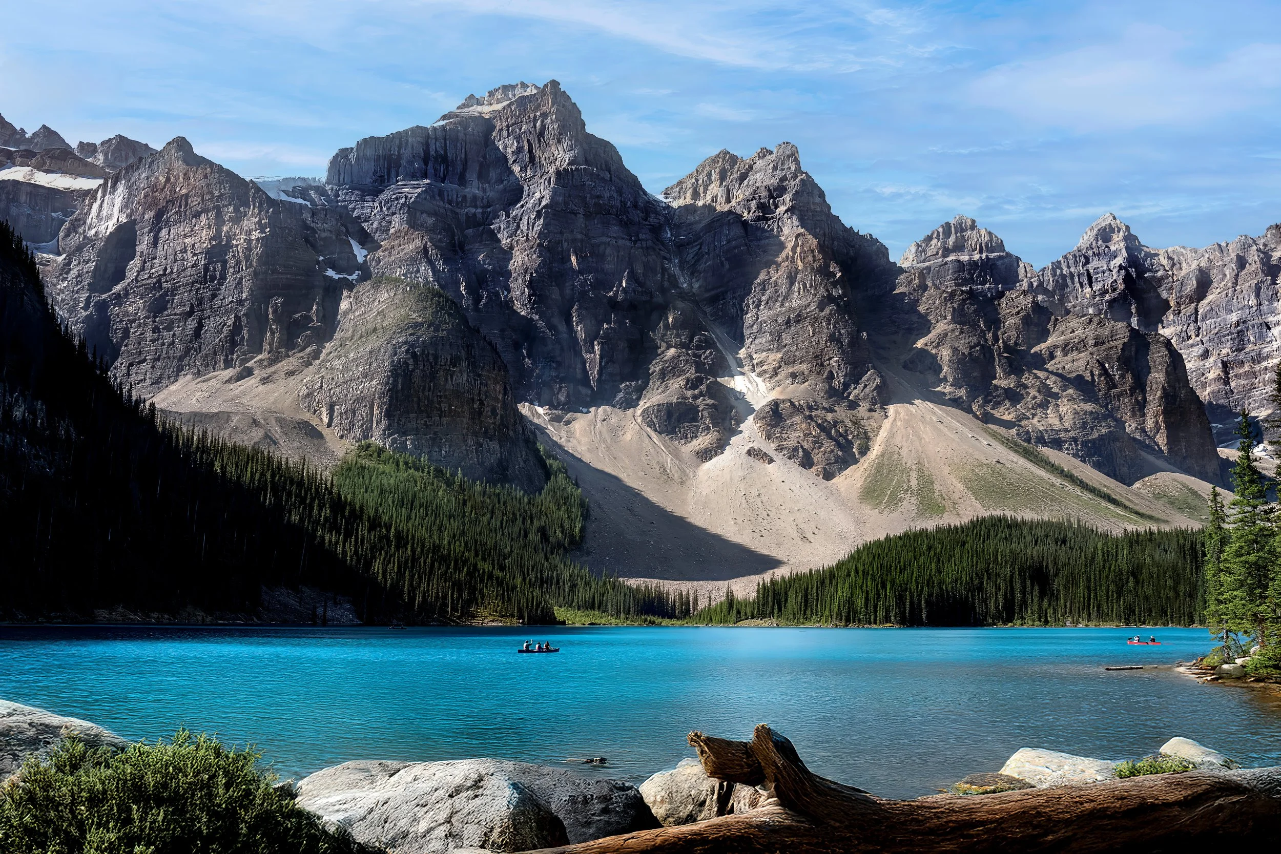 A mountain range with rugged peaks and patches of snow, a turquoise lake in the foreground, and a forested shoreline with green trees. There are small boats on the lake with people paddling.
