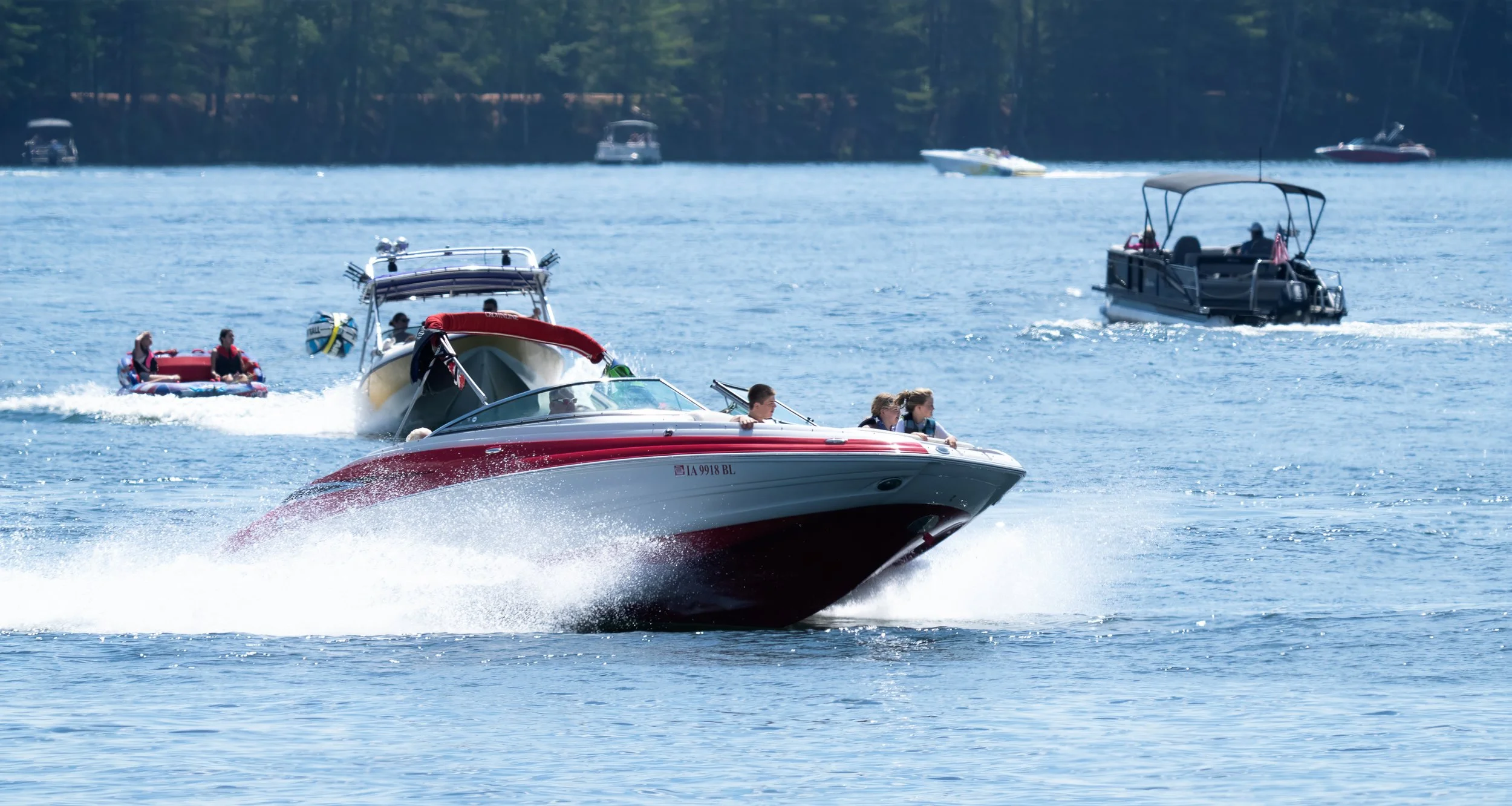 Several boats and speedboats on a lake with people enjoying a day of boating and water activities, with trees on the shoreline in the background.