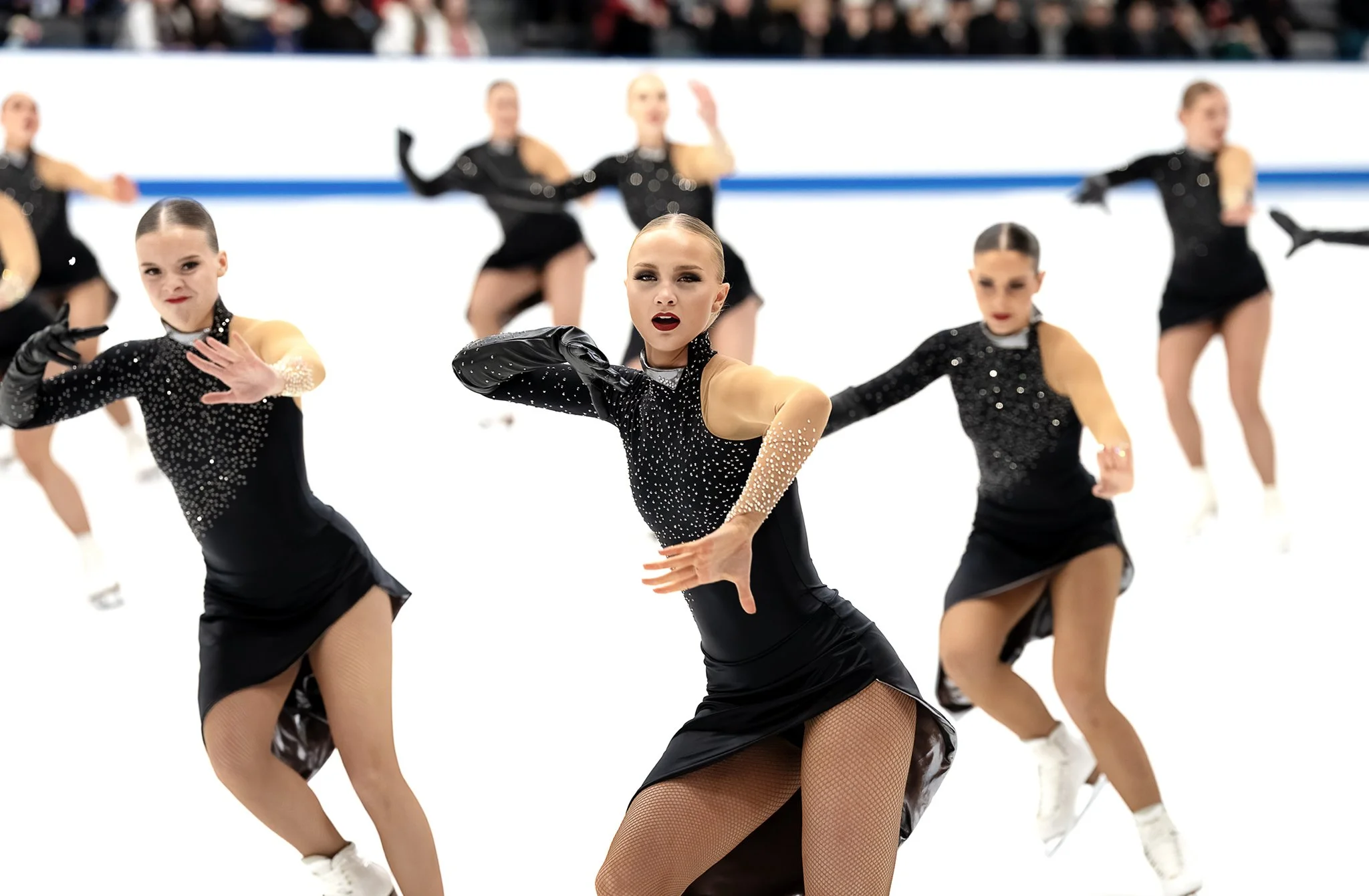 A group of female figure skaters in black costumes with rhinestones performing a synchronized routine on ice.