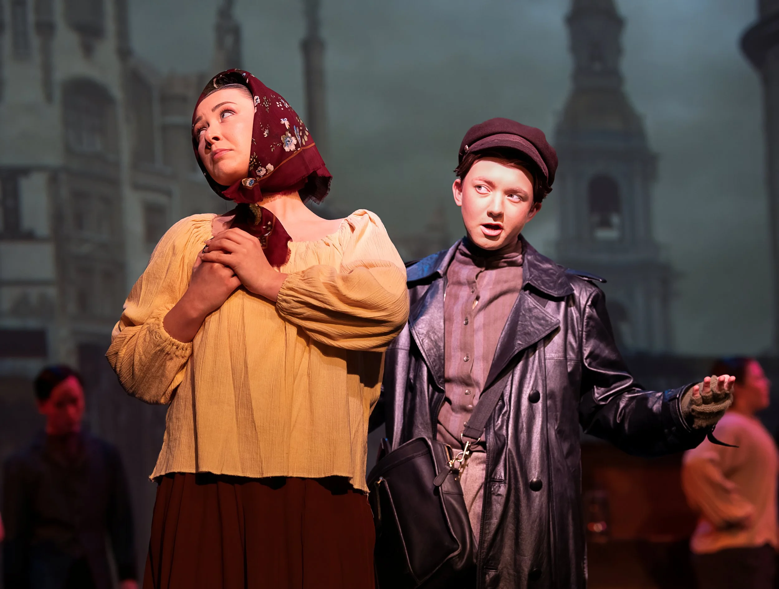 People dressed in period costumes stand on stage during a theater performance with a dramatic background of a building facade.