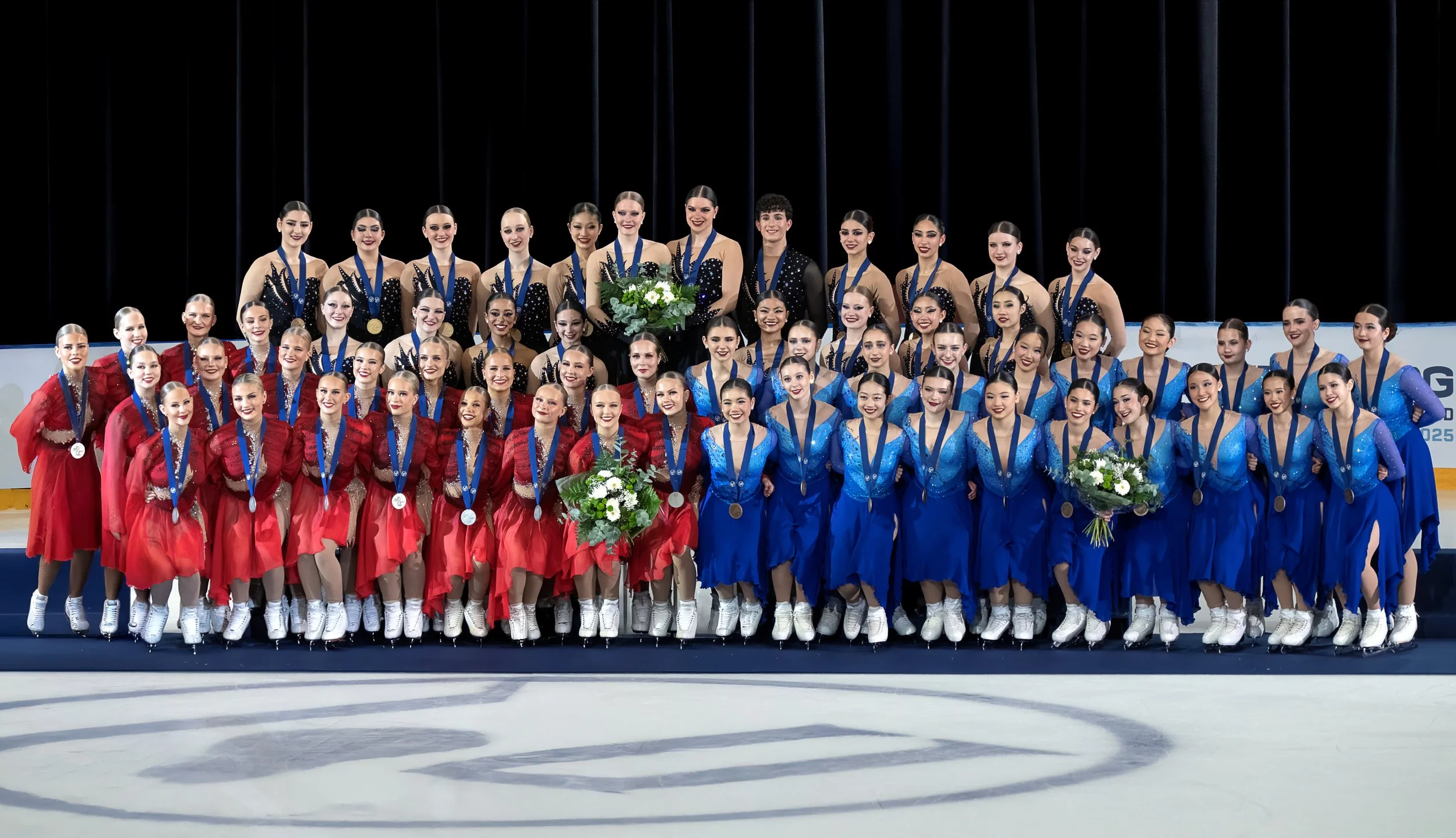 Group of figure skaters in colorful costumes standing on ice rink, wearing medals, some holding bouquets, with dark background.