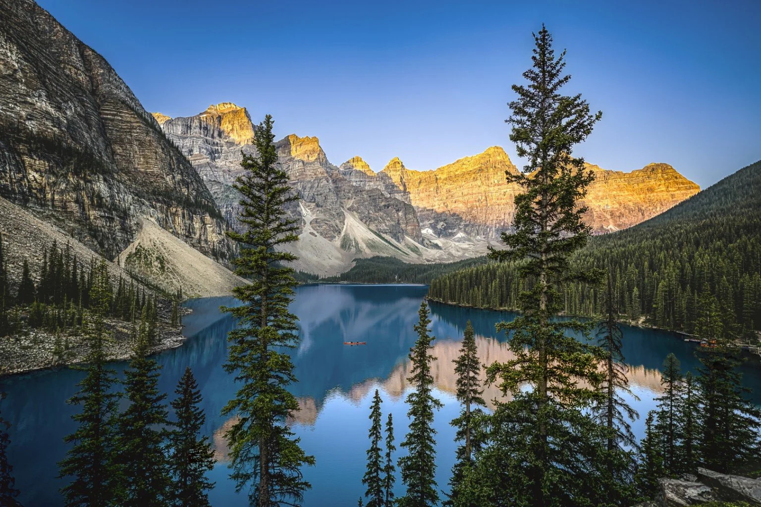 A scenic view of a mountain lake with green pine trees along the shoreline, surrounded by tall rugged mountains with snow patches, under a clear blue sky, during sunset.